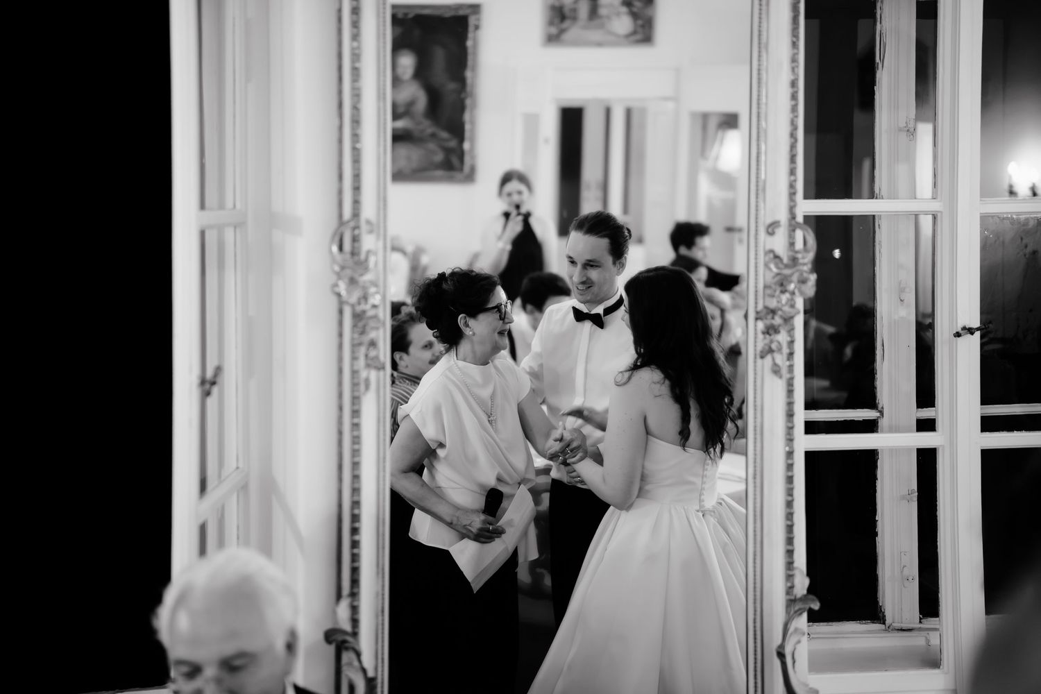 Black and white wedding photo of bride and groom sharing a moment with guests in an elegant venue.