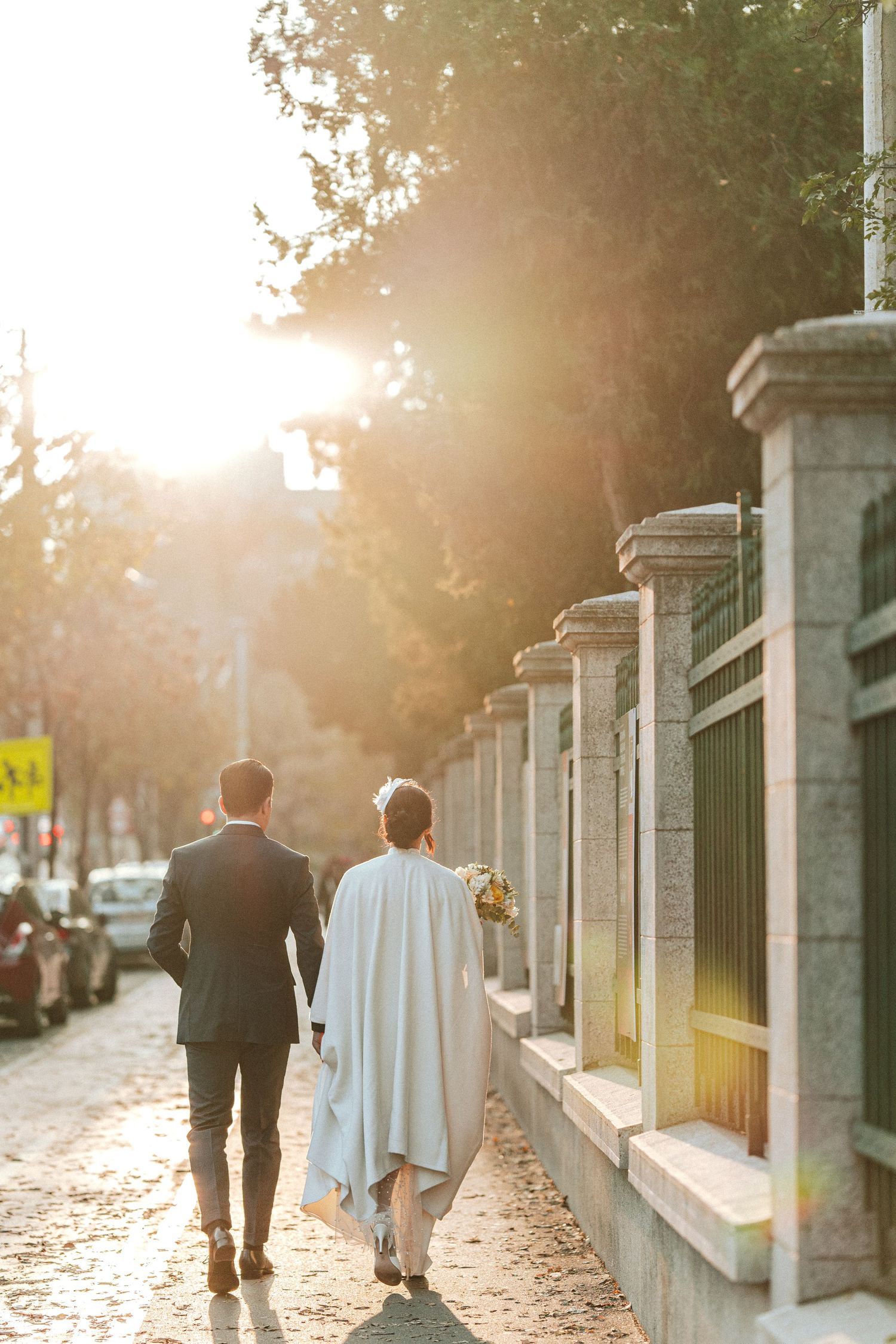 Wedding couple walks along stone wall path at sunset creating silhouettes.