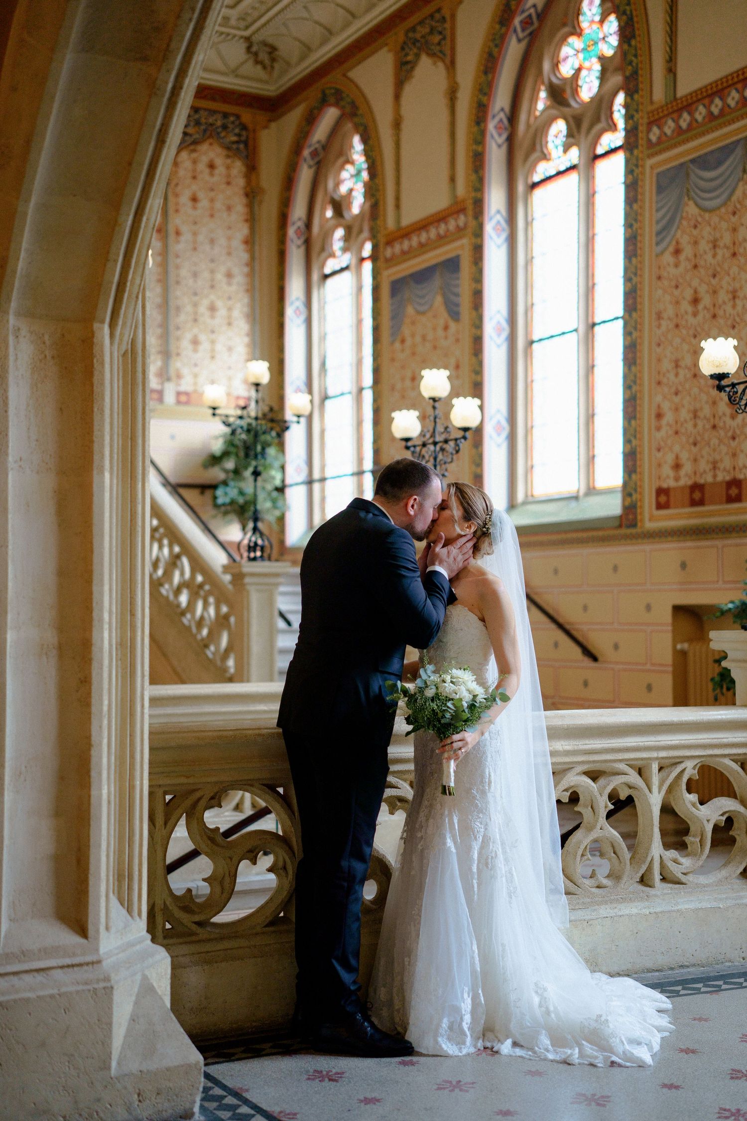A romantic wedding moment captured on an elegant staircase with gothic windows in the background.
