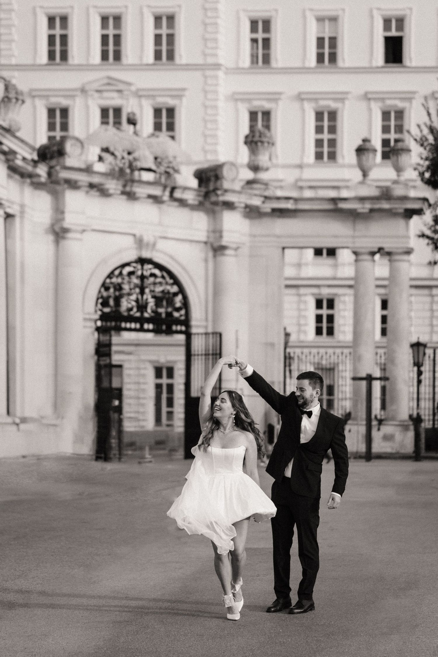 A black and white wedding photo sequence showing a couple dancing playfully in front of an elegant historic building.