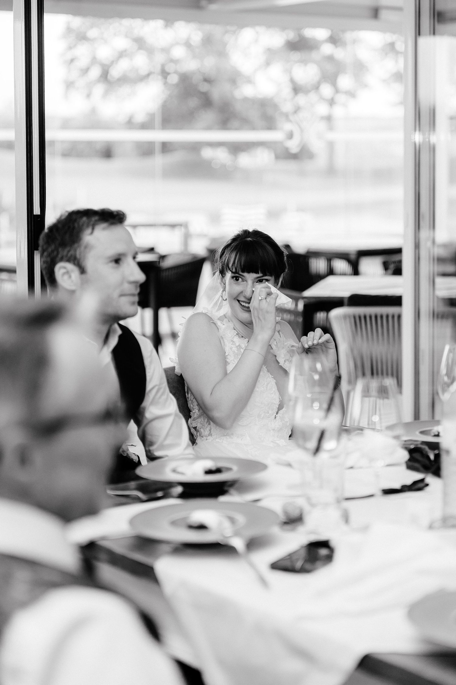 Black and white photo of an emotional bride wiping tears at wedding reception dinner table with groom beside her.