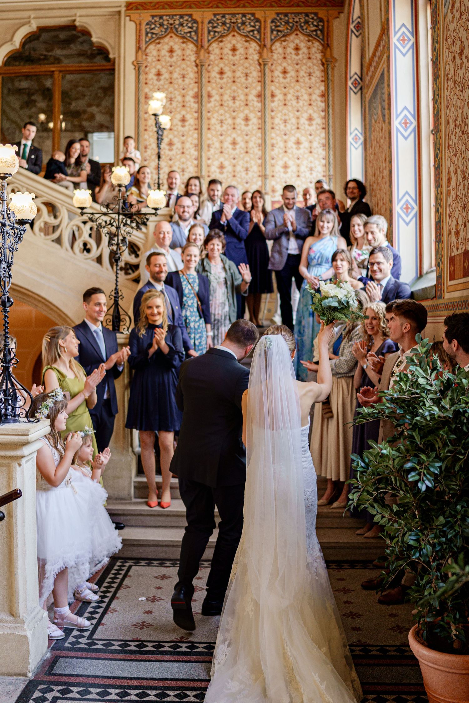 Wedding party and guests gather in an elegant Victorian hallway for celebration photos.