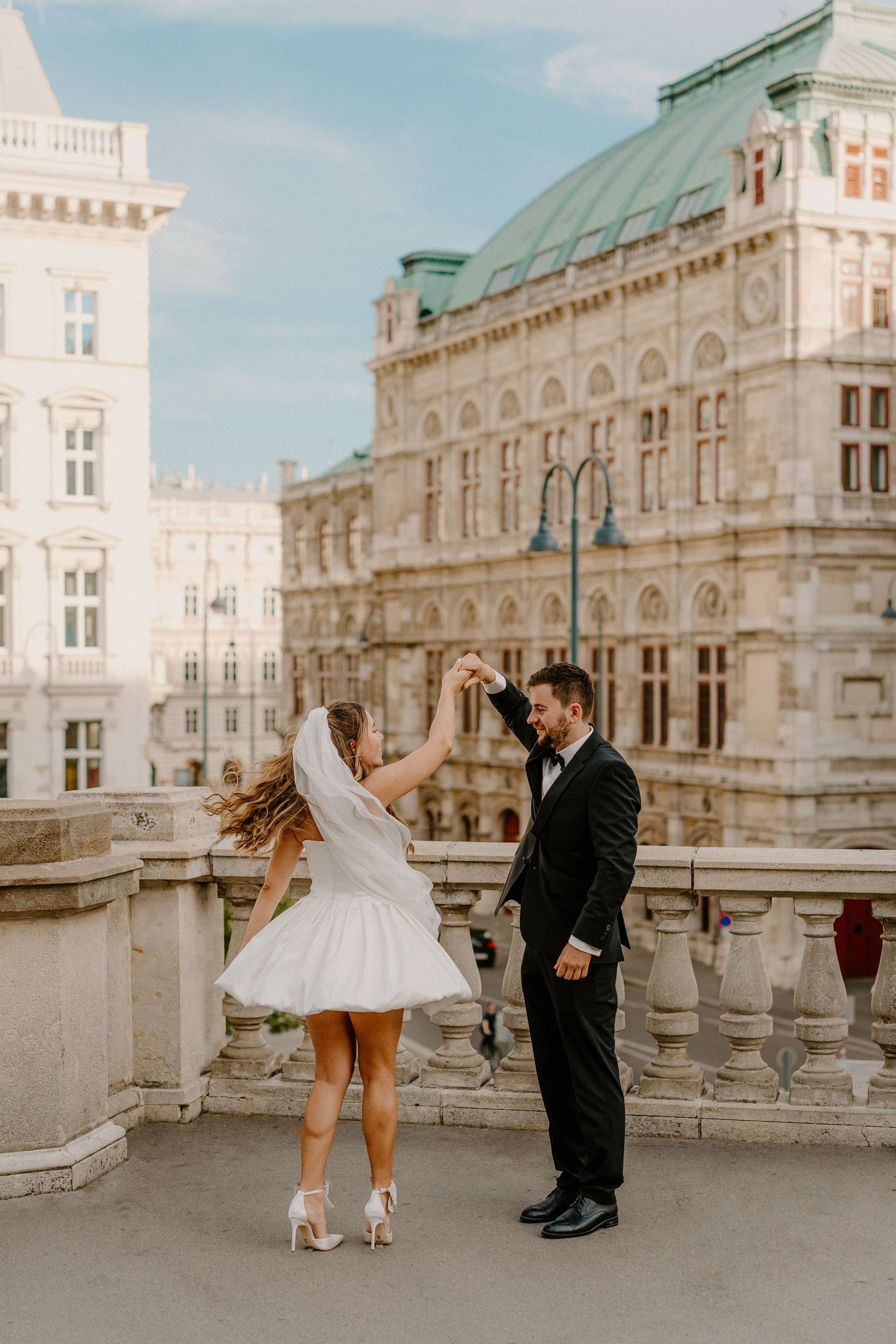 A couple in wedding attire dancing on a balcony at the Vienna State Opera House with ornate architecture in background.
