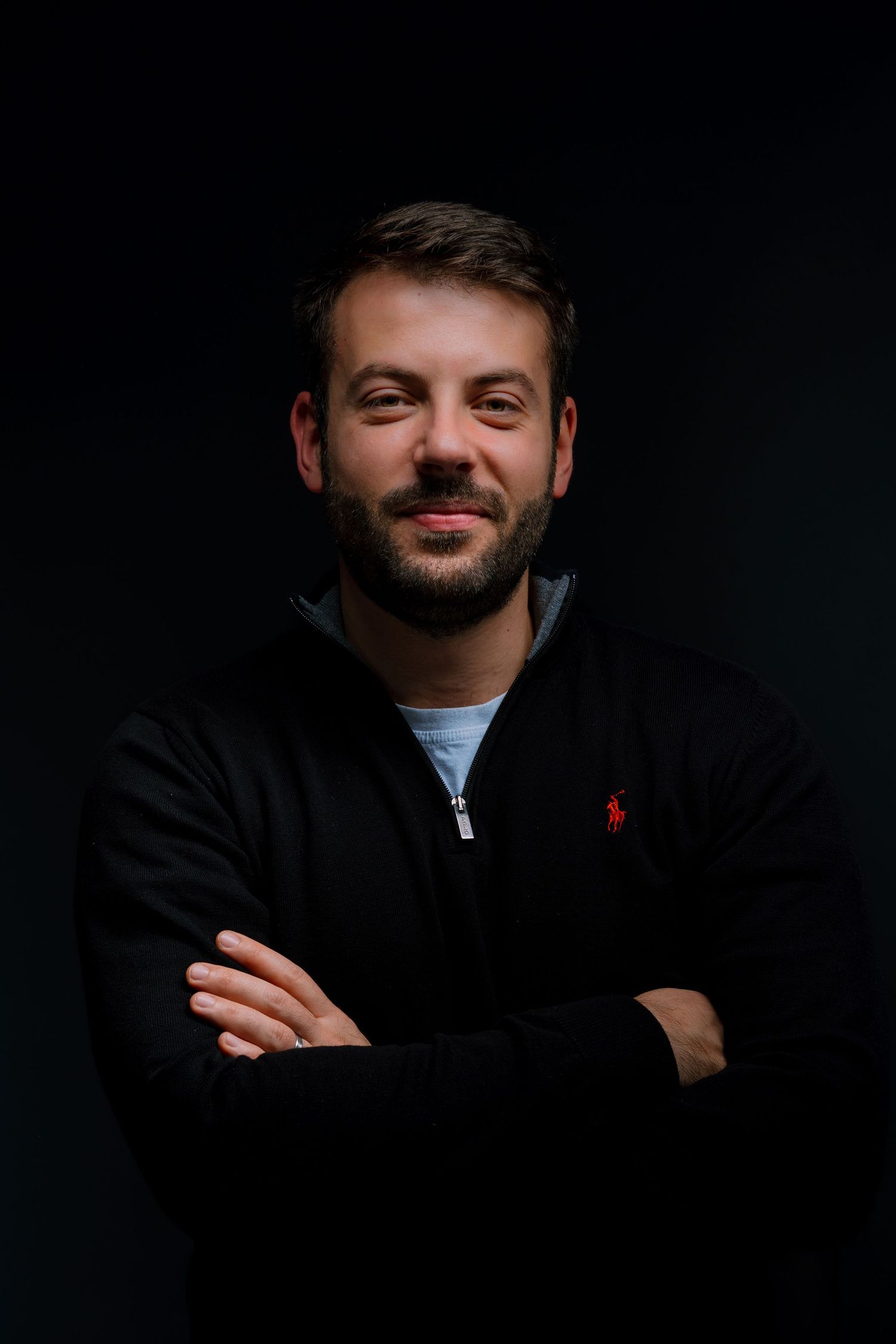 Man with beard and crossed arms smiling, wearing black Ralph Lauren zip-up hoodie against dark background.