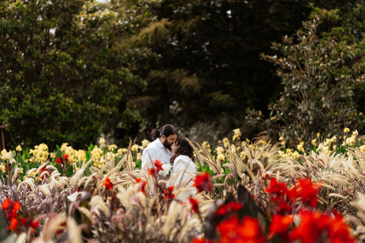 Couple embracing romantically among vibrant red and golden flowers in a lush garden setting.