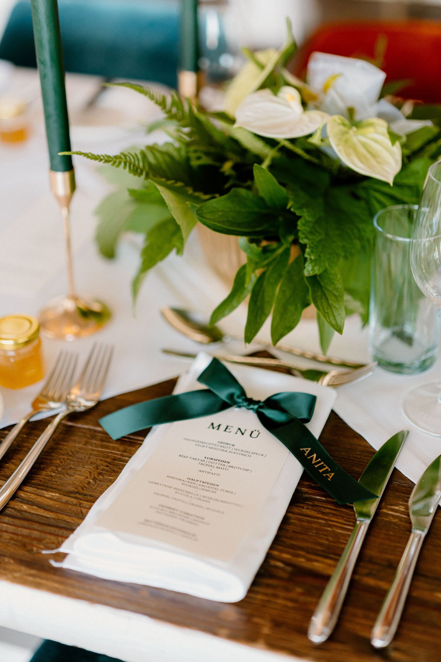 Elegant wedding table setting with green floral centerpiece, gold cutlery, and a menu card tied with a dark green ribbon.