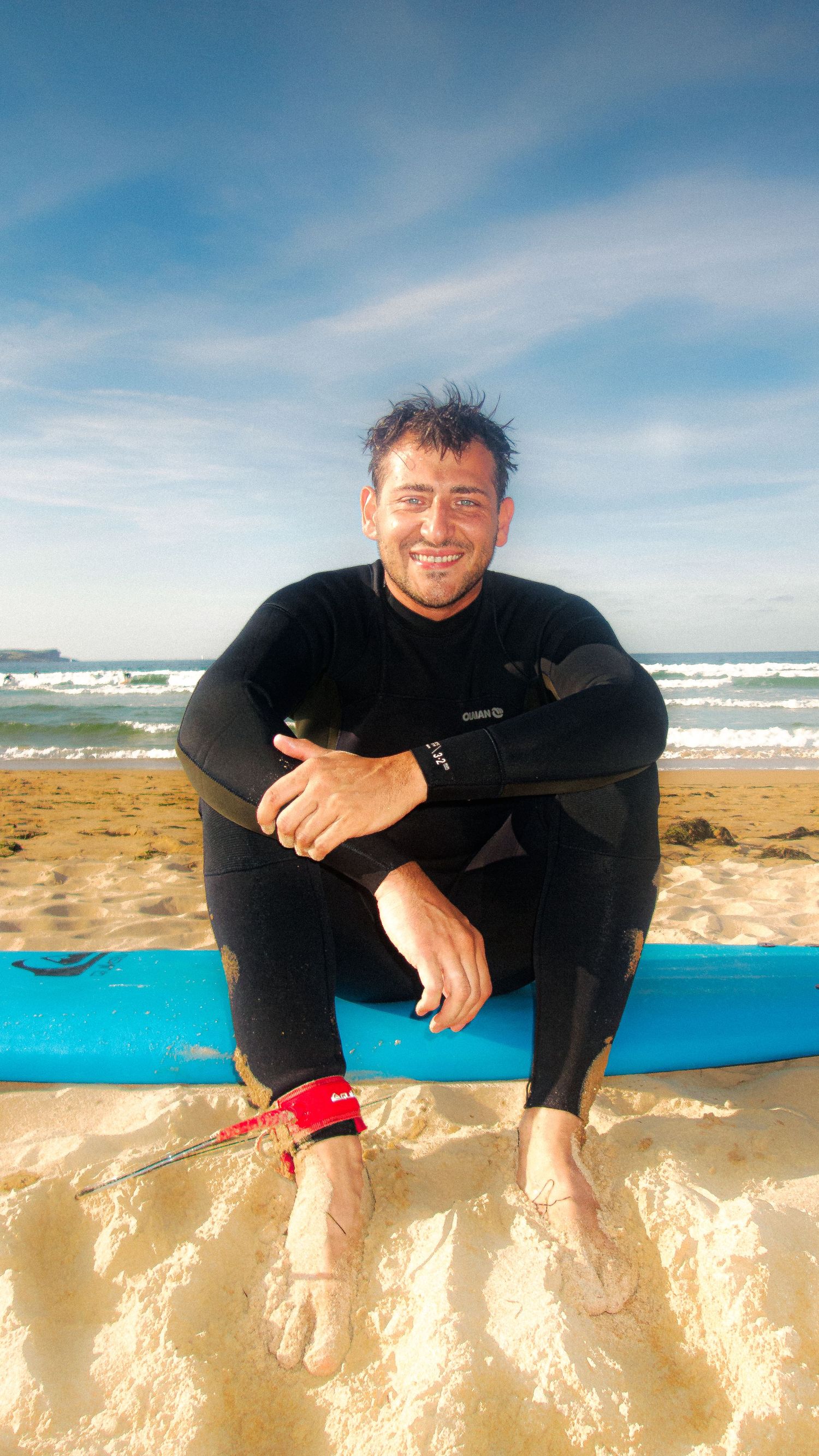 Person in wetsuit relaxing on sandy beach with blue surfboard and ocean waves in background.