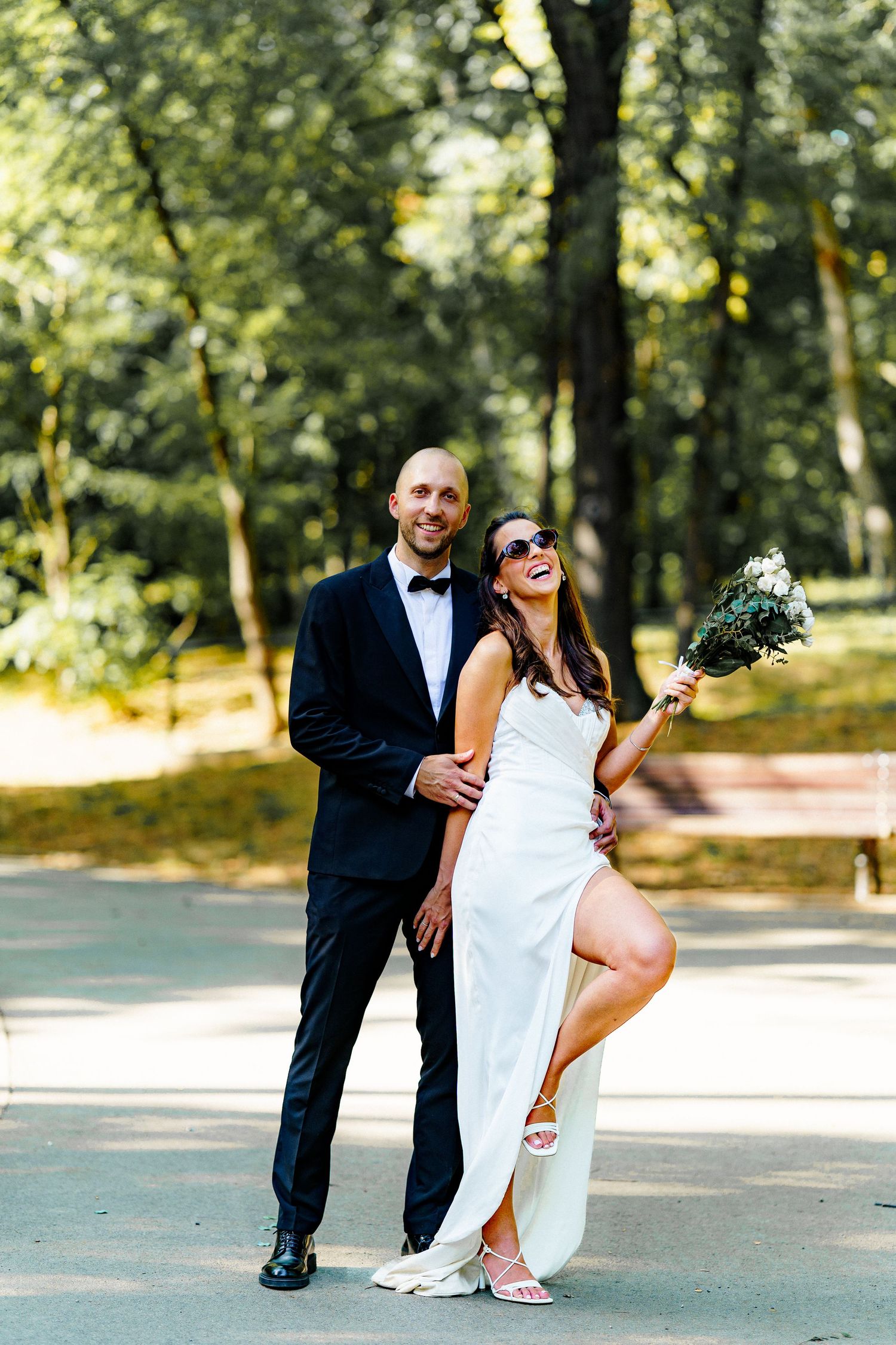 Newlywed couple shares playful moments on a tree-lined path during their wedding photoshoot.