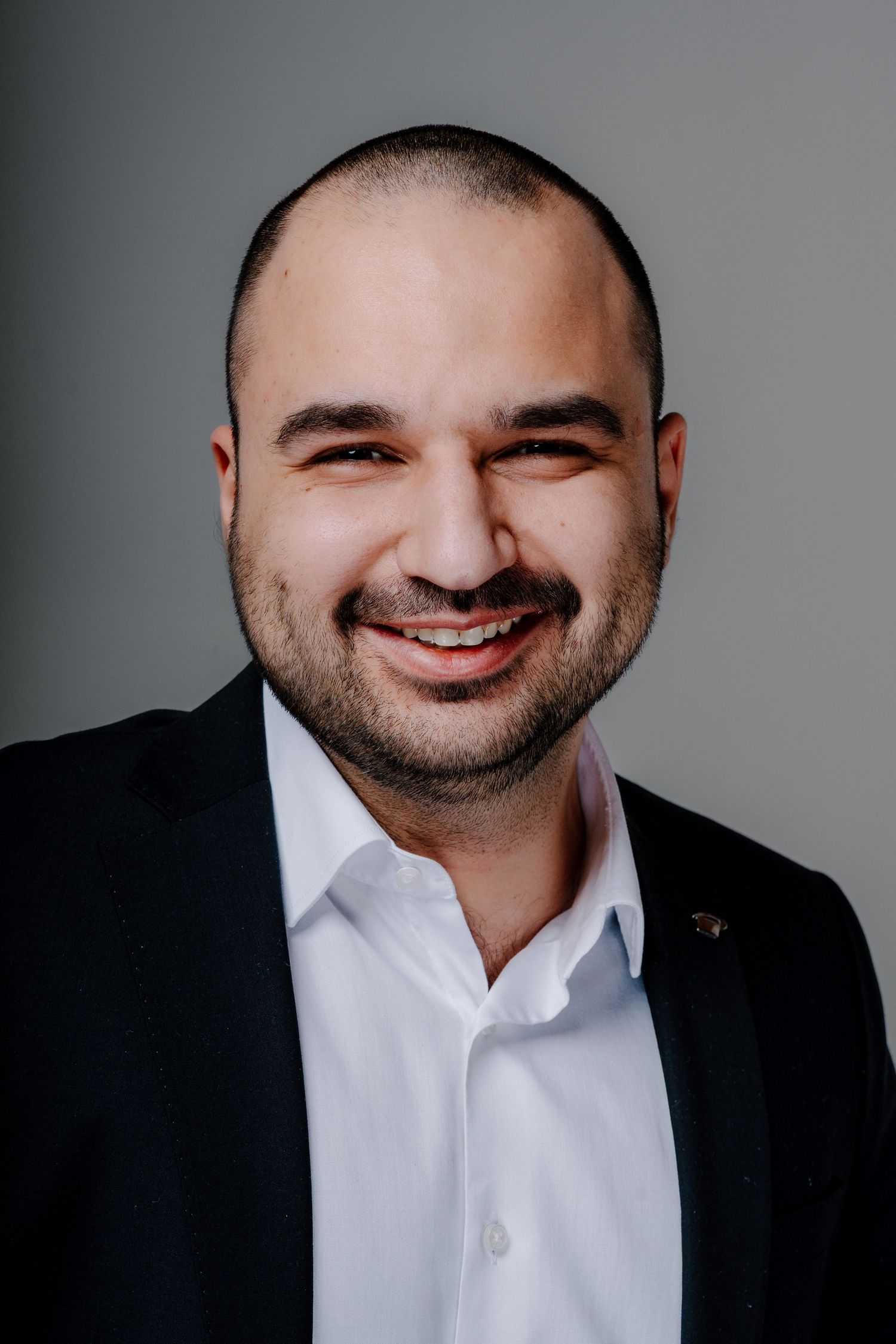 Smiling man in black blazer and white shirt with short dark hair and beard poses for professional headshot.