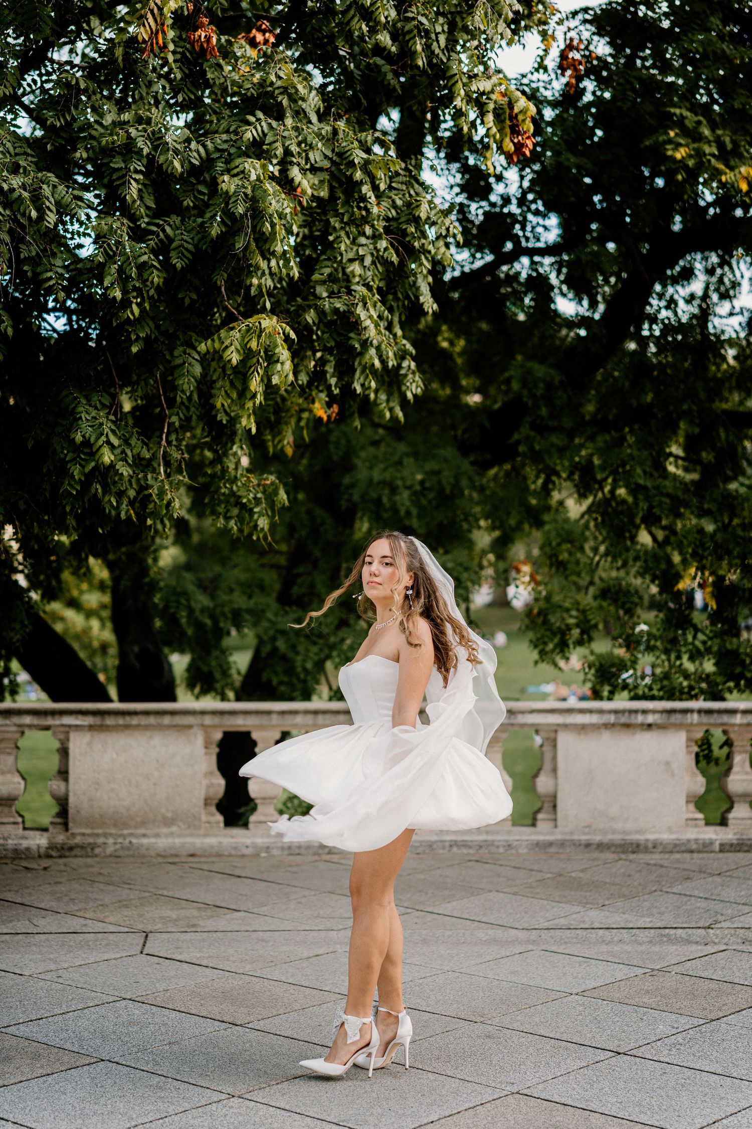 A bride in a flowing white dress twirls gracefully on a stone terrace surrounded by green trees.