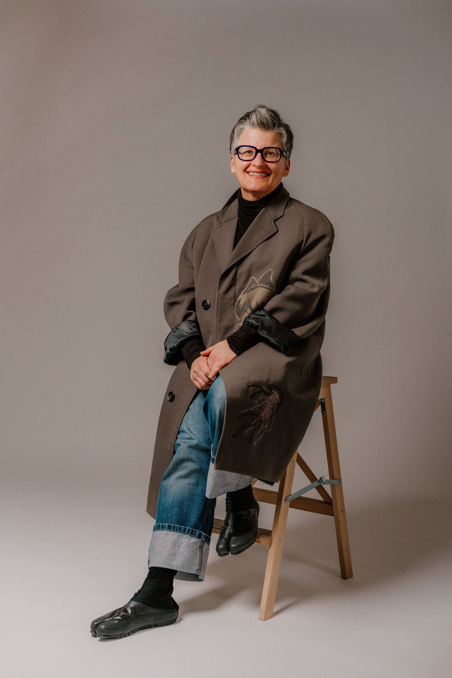 Older woman with gray hair and glasses sits on a wooden stool wearing an oversized coat and cuffed jeans.