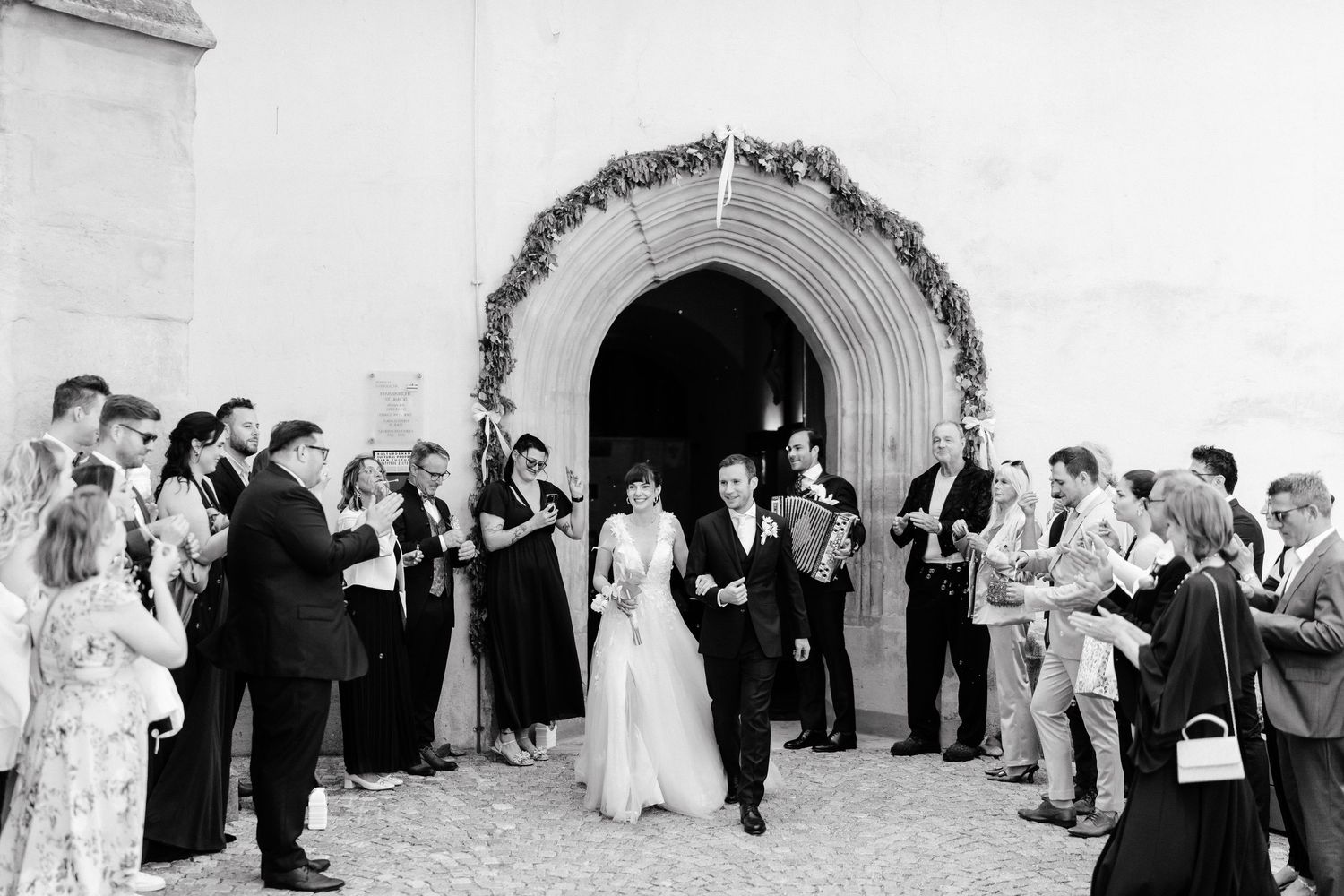 Bride and groom exit church through floral arch, greeted by cheering guests in elegant attire at outdoor wedding ceremony.