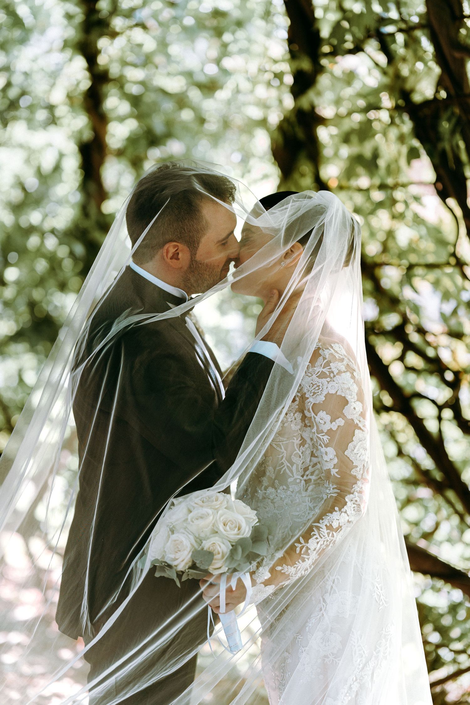 A tender wedding kiss captured through a flowing white veil in a natural outdoor setting.