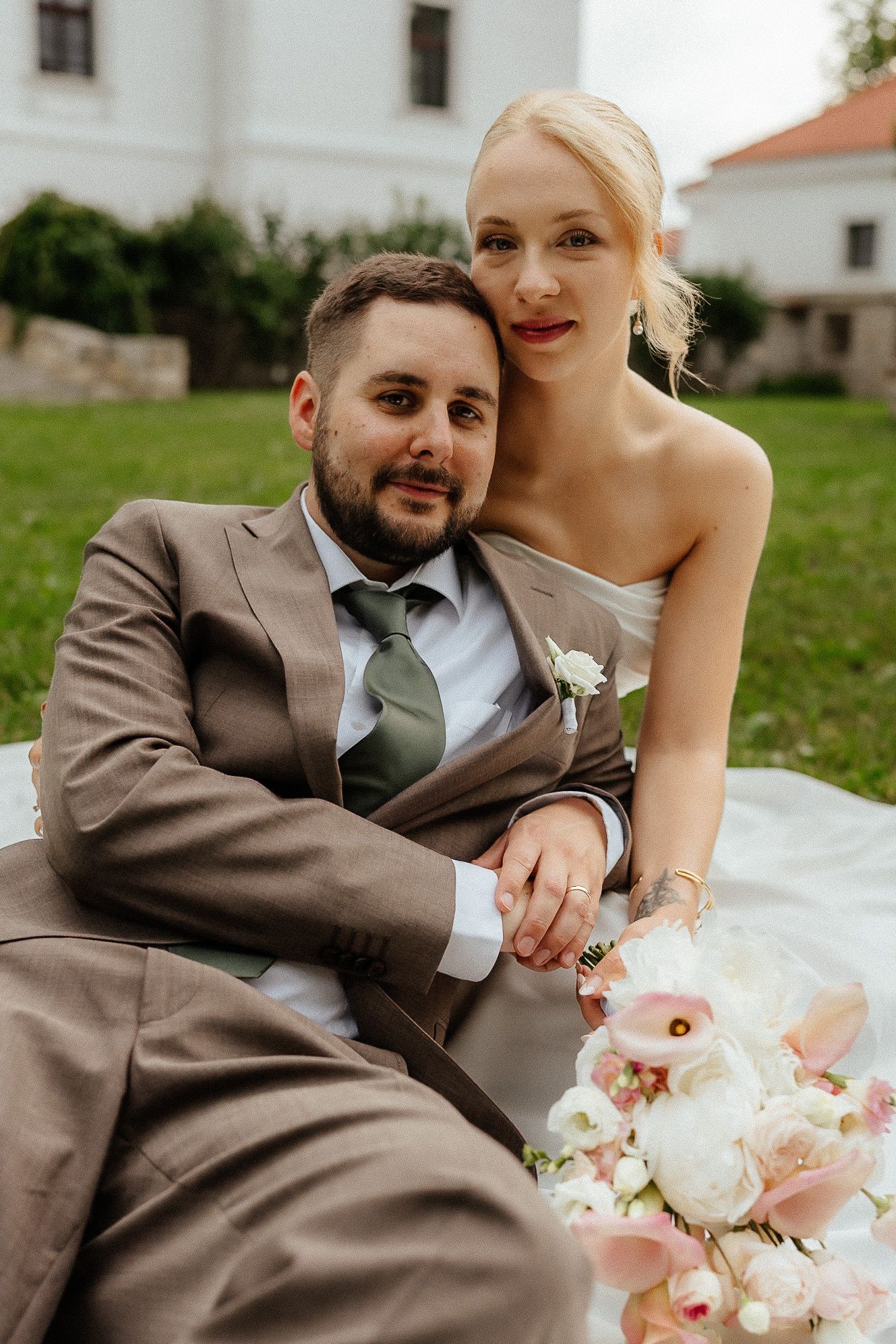 Bride and groom sitting on grass, groom in brown suit with green tie, bride in white dress holding pink and white bouquet.