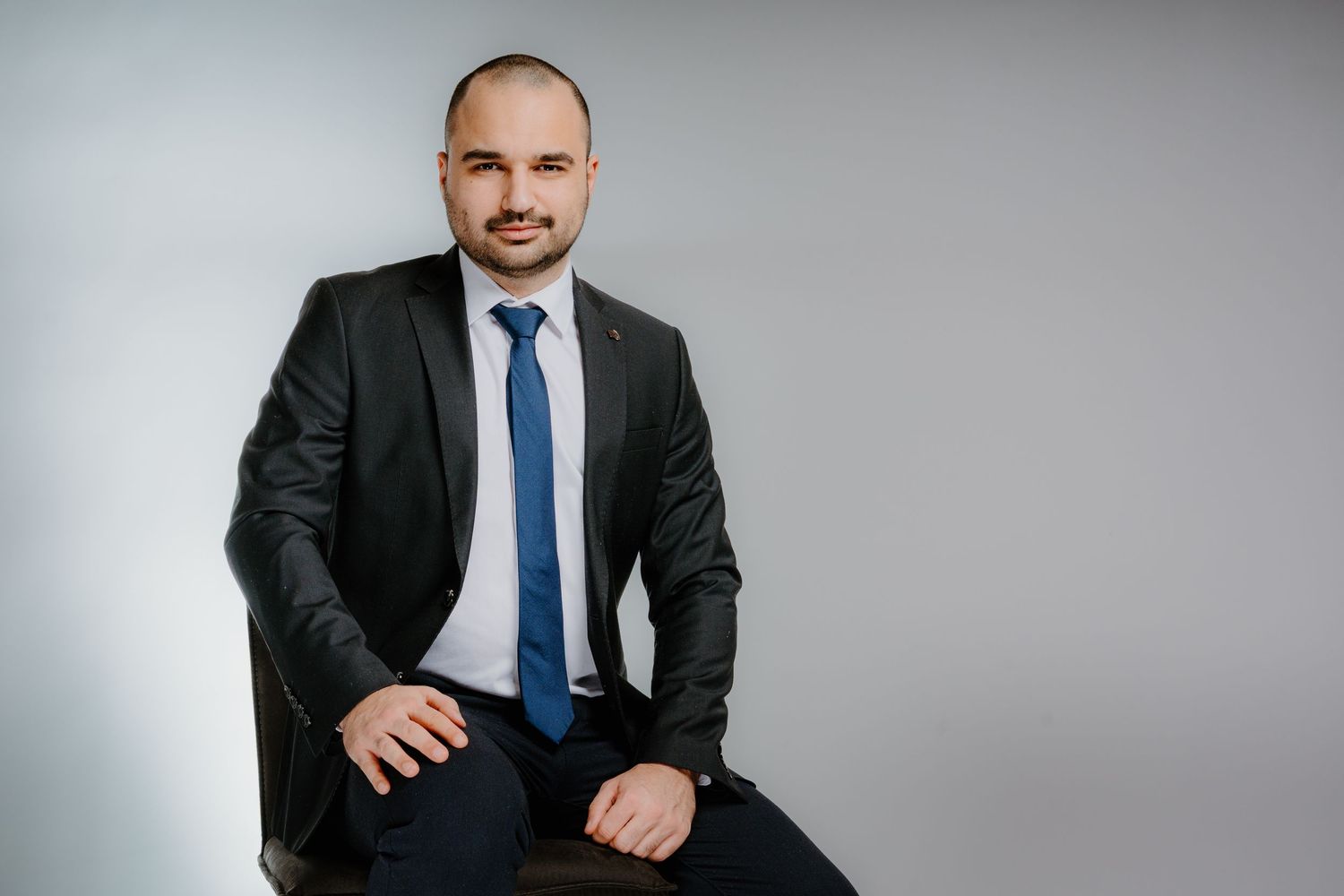 Professional businessman in dark suit and blue tie sitting confidently against a light gray background.