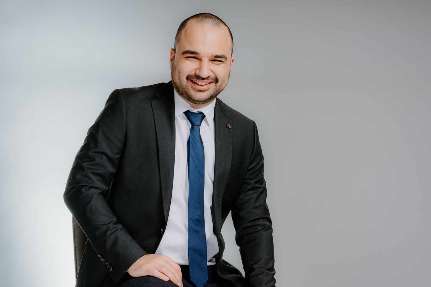 Professional businessman in black suit and blue tie smiling confidently while seated against a light gray background.
