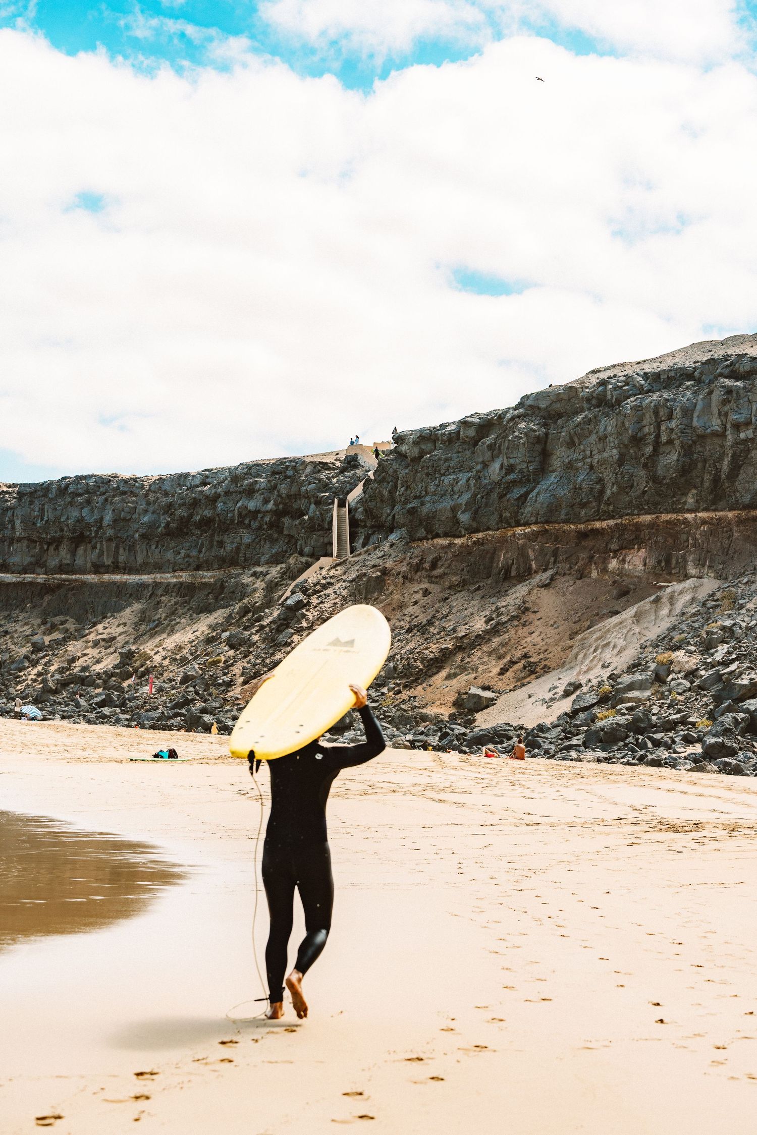 Someone walking along a sandy beach carrying a yellow surfboard with cliffs in the background.