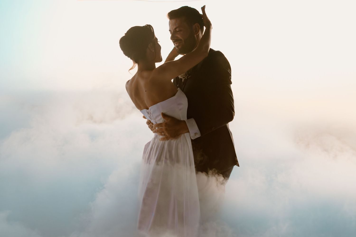 Dreamy wedding couple embracing against soft cloudy sky.