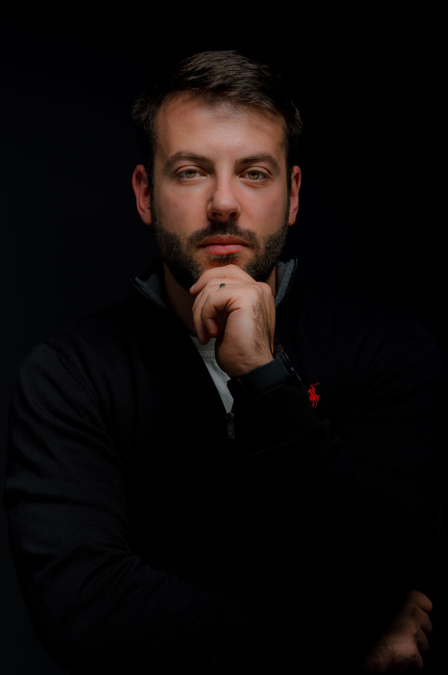 Man with beard in dark studio portrait, hand on chin, wearing black jacket, thoughtful expression.