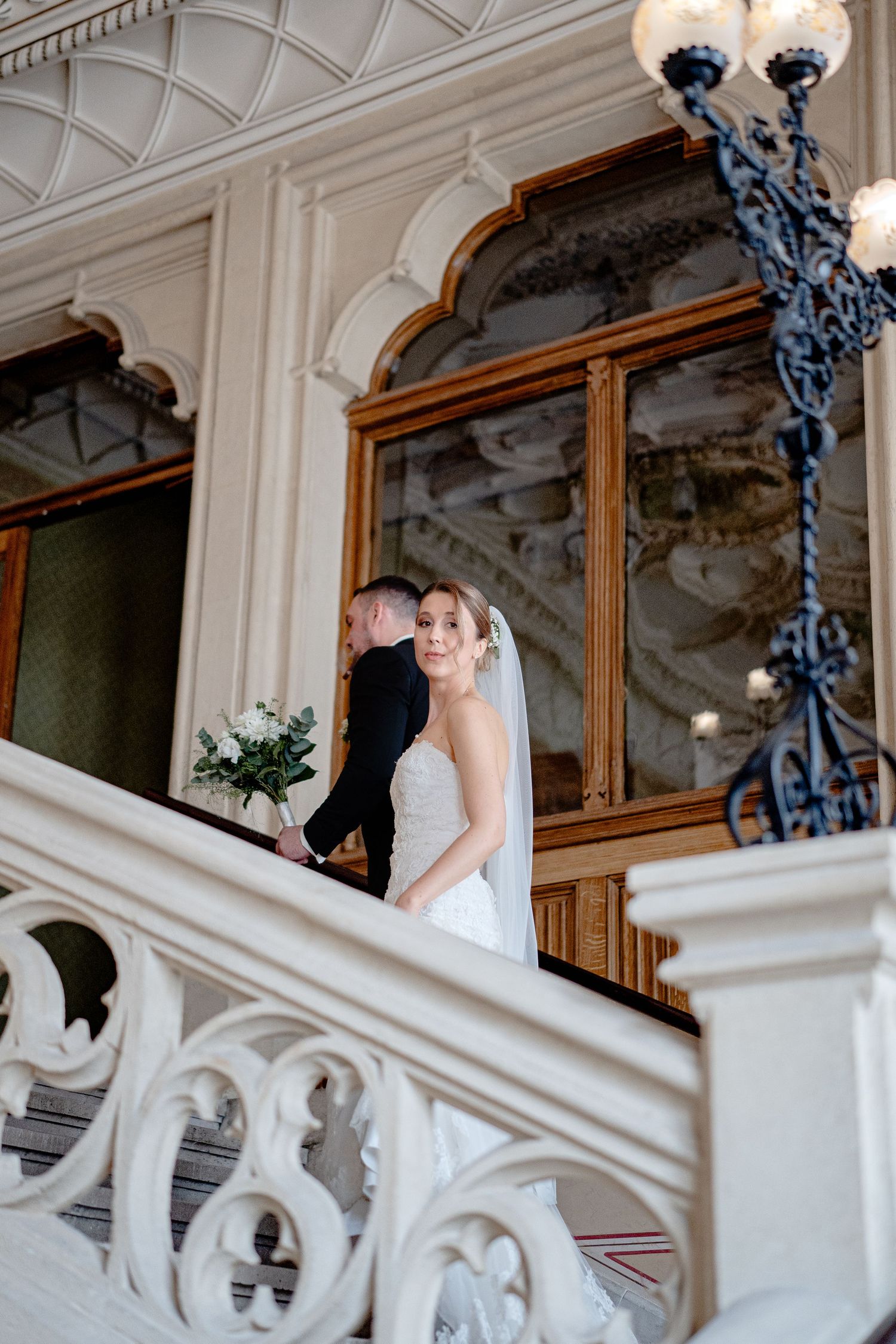 A bride stands on an ornate white staircase with decorative iron lighting fixtures.