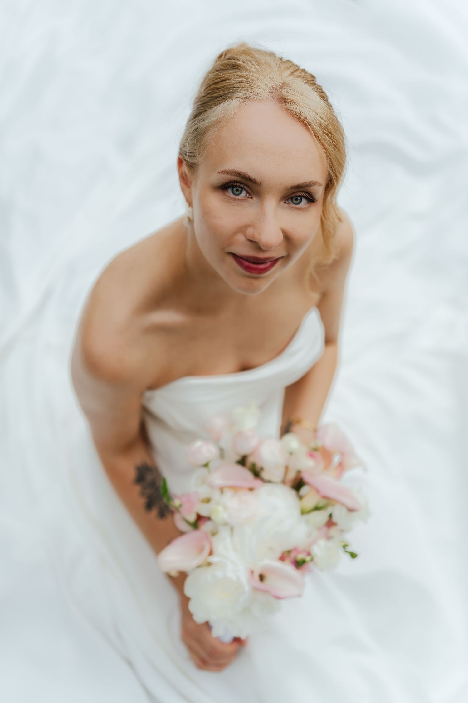 Blonde bride in strapless white gown holding pink and white bouquet, smiling up at camera with red lips.