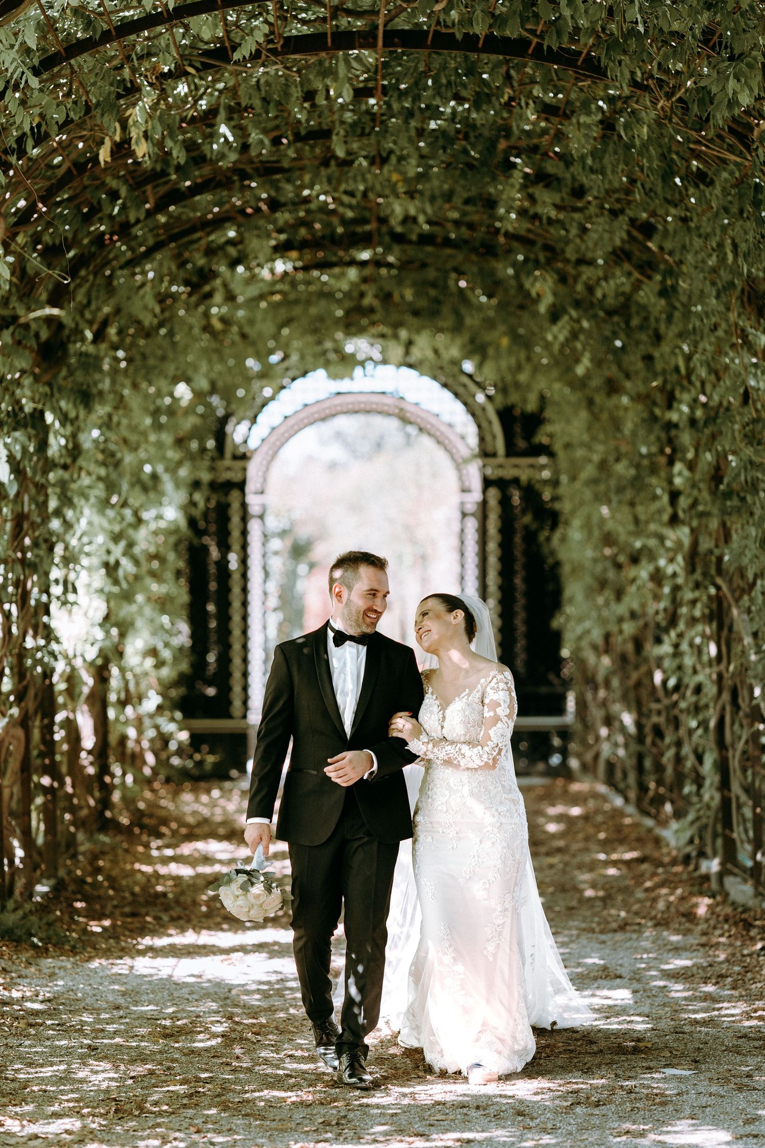 A romantic wedding photo of a couple walking through a lush garden archway in formal attire.