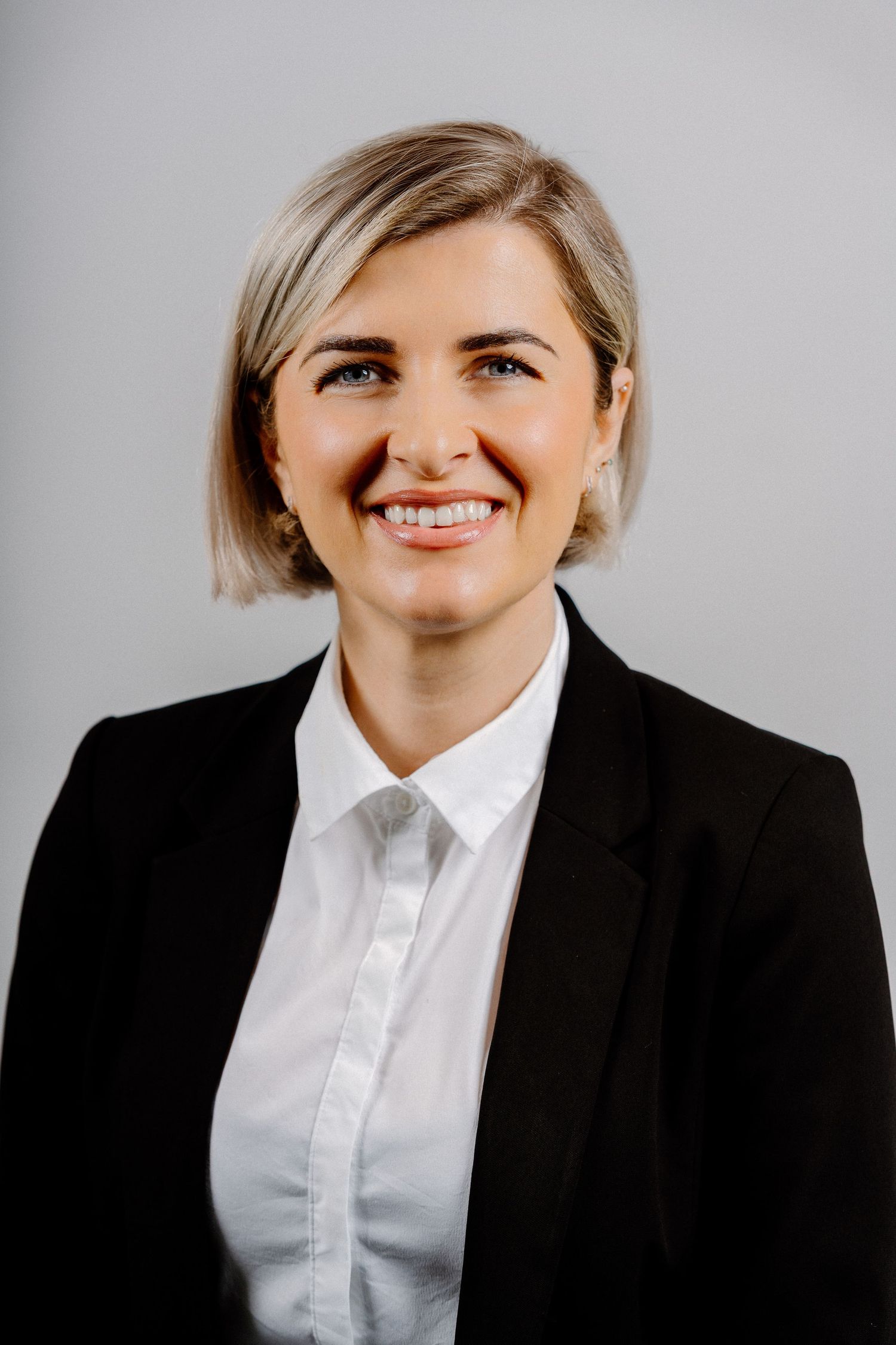 Professional woman in black blazer and white shirt smiling confidently against a gray background.