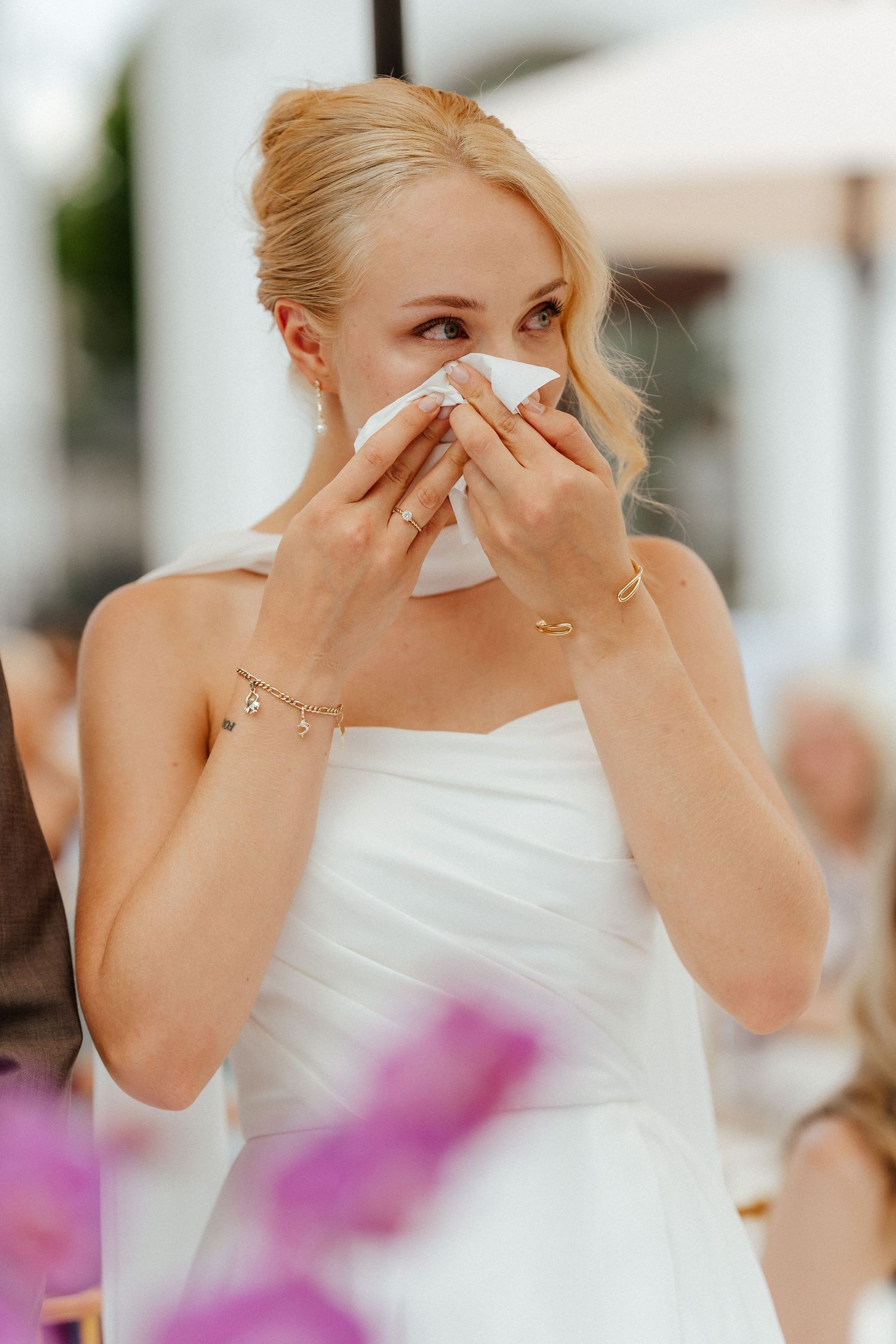 Emotional bride in white strapless gown dabbing tears with tissue, wearing gold jewelry at outdoor wedding.