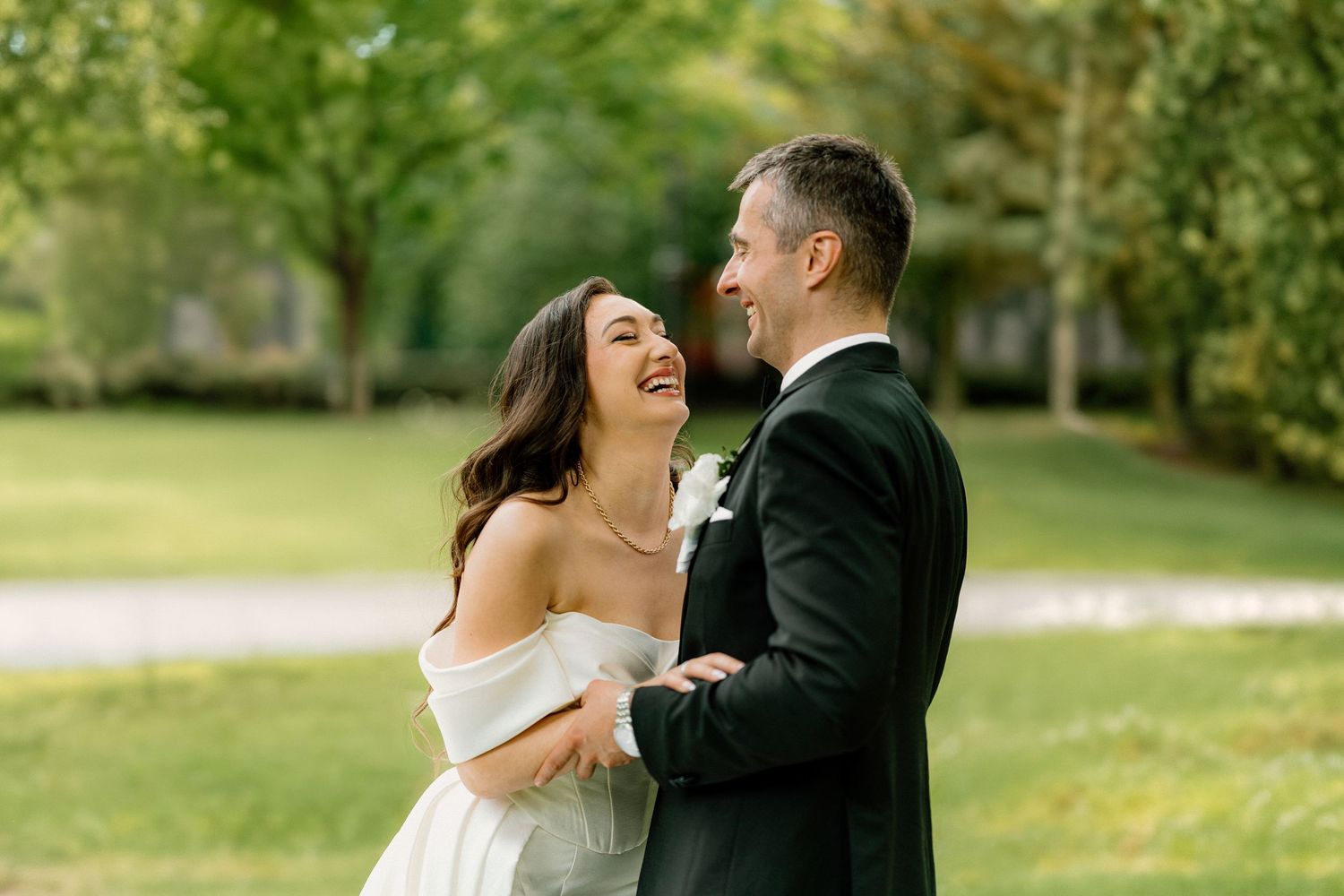 A couple shares joyful laughter together during an outdoor wedding photoshoot in a lush green garden setting.