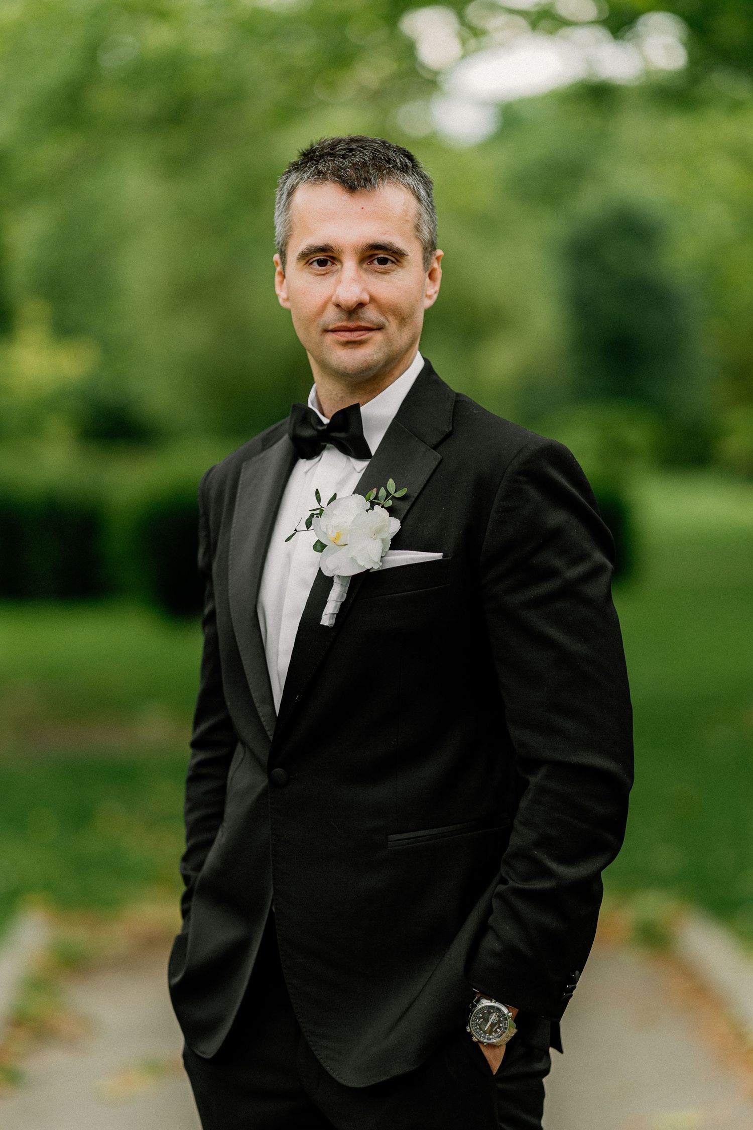 Groom in classic black tuxedo poses for formal portraits in garden setting.