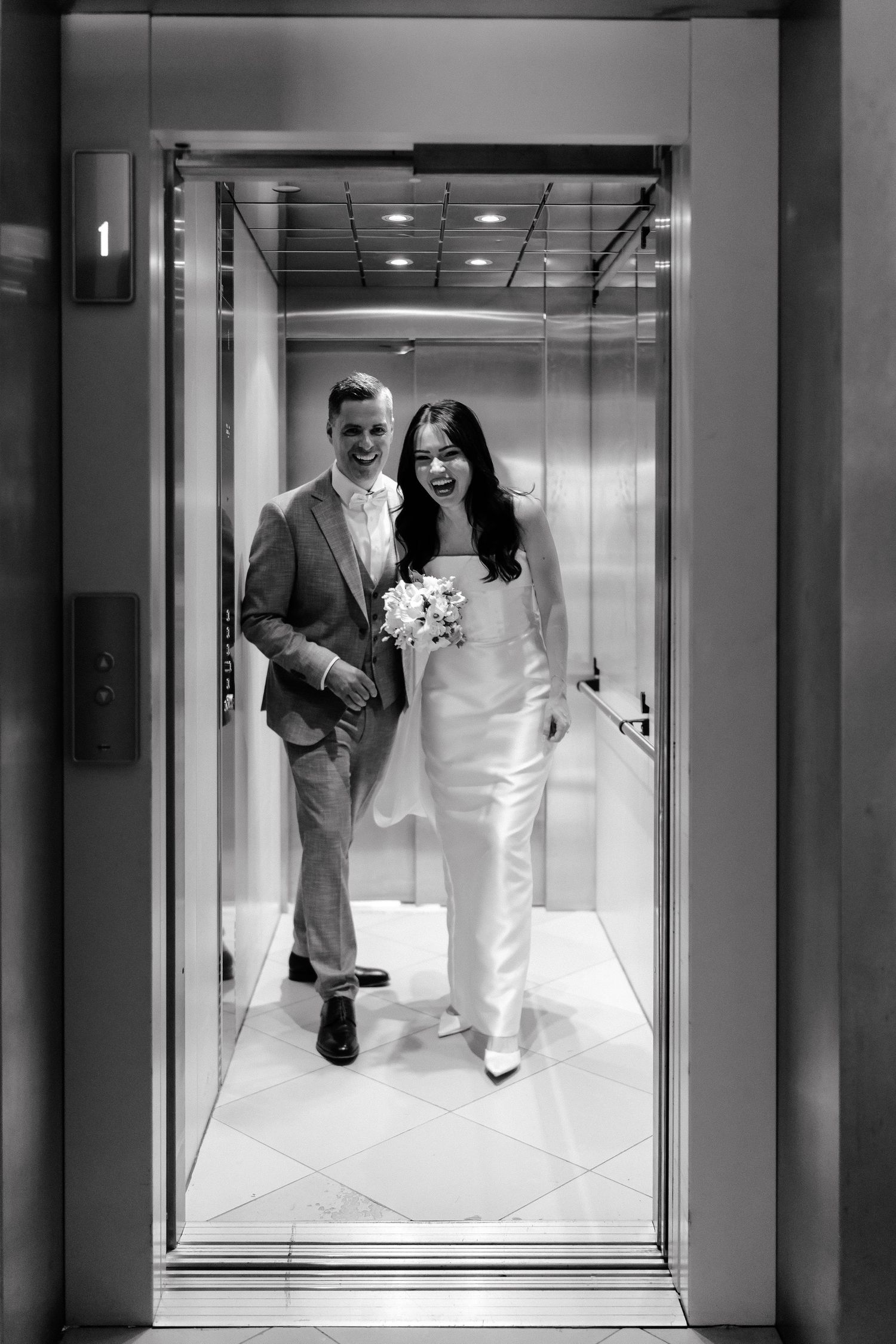 Black and white photo of a smiling bride holding bouquet and groom stepping out of an elevator on their wedding day.