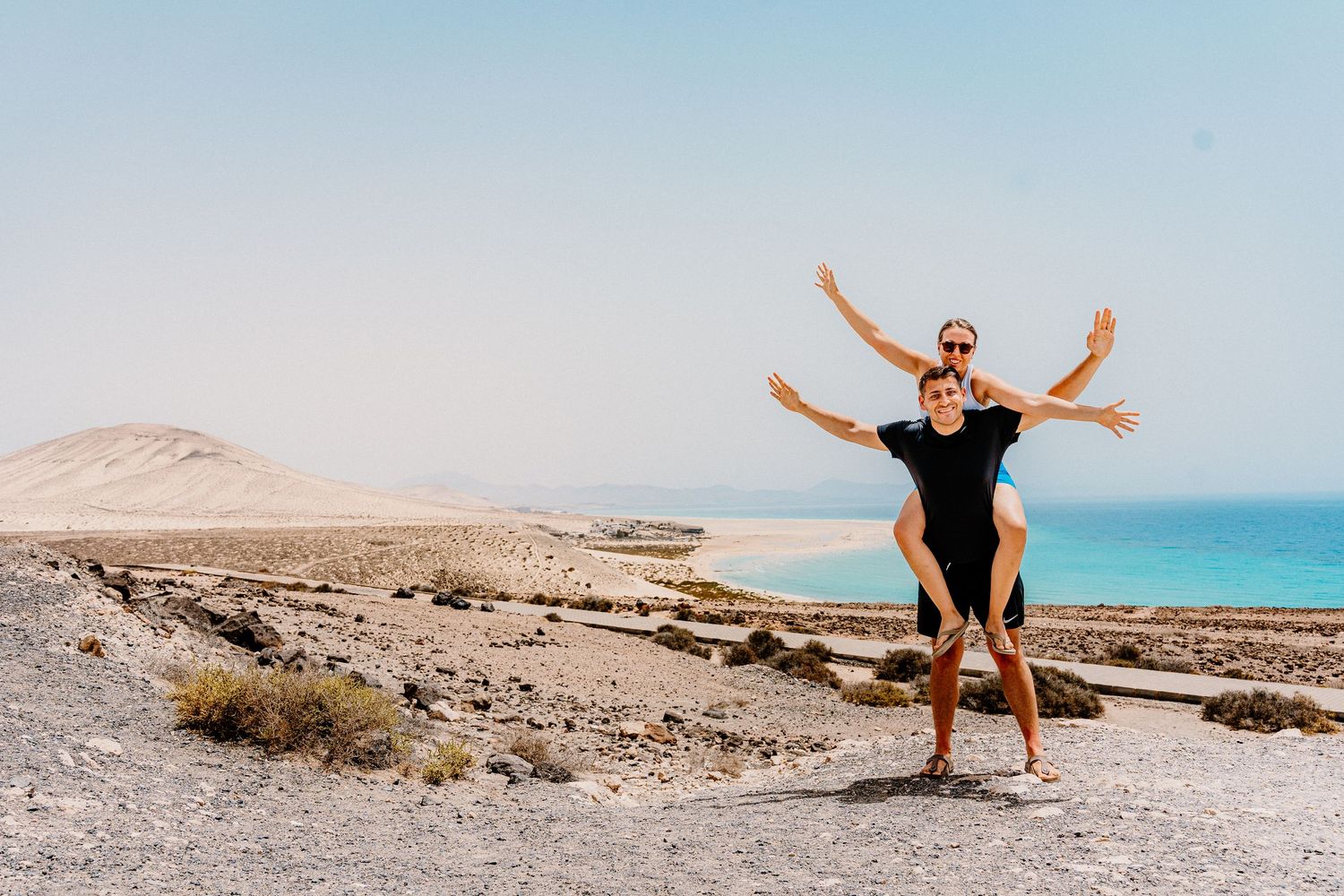 A person poses with outstretched arms on a desert cliff overlooking the ocean.