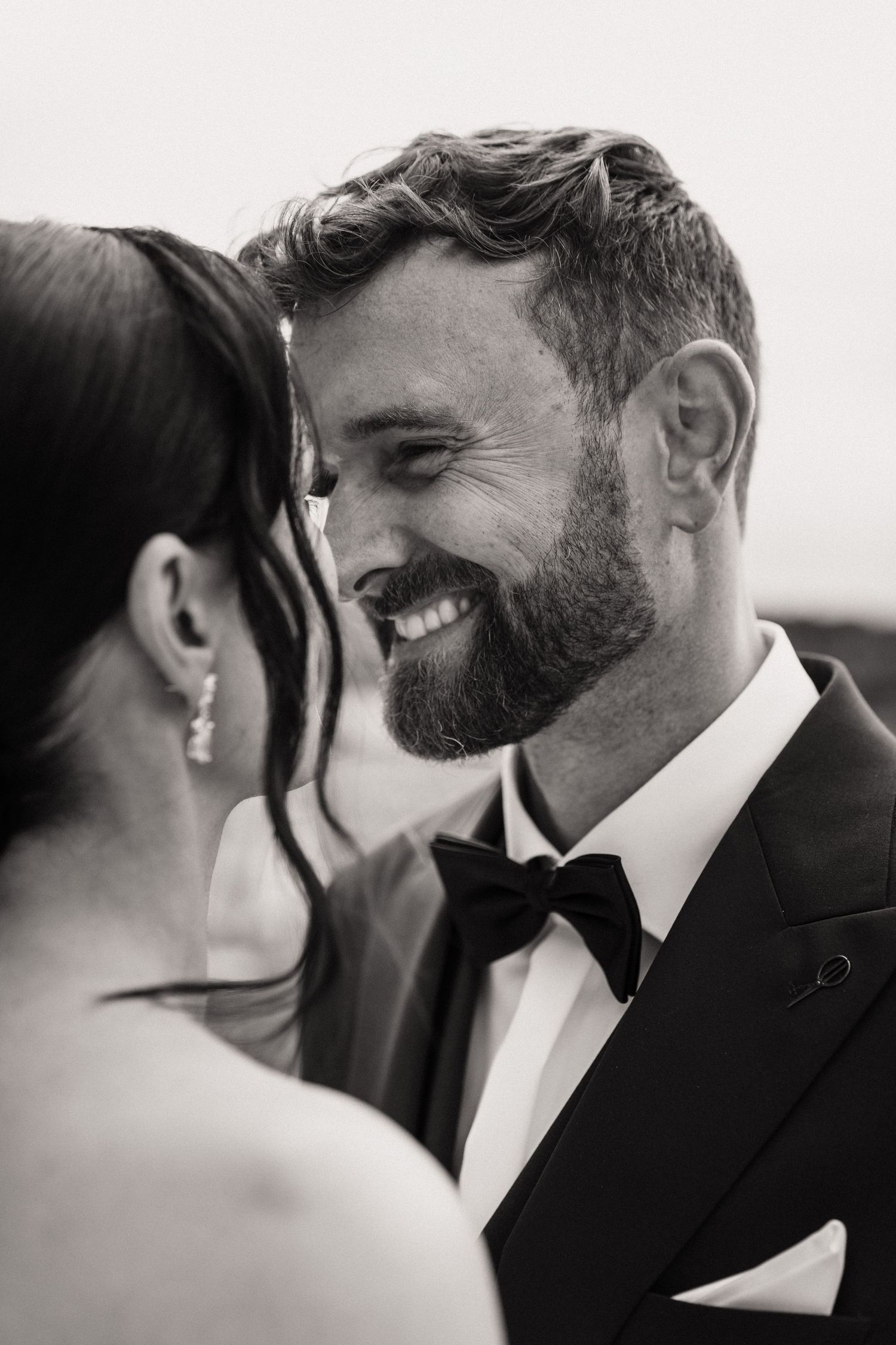 Black and white photo of a smiling bearded groom in a tuxedo sharing an intimate moment with his bride.