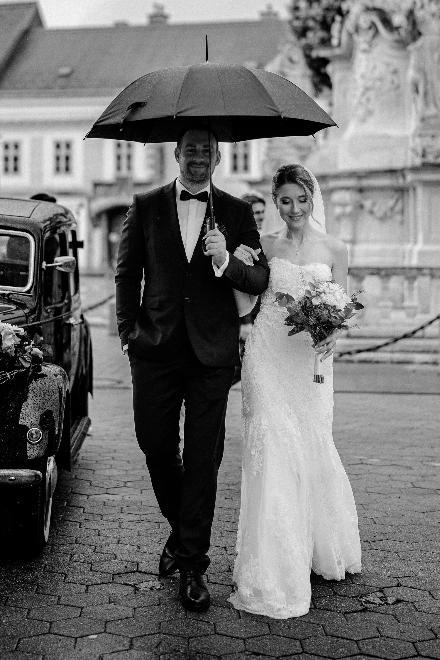 A couple walks on cobblestone streets in formal wedding attire with a black umbrella on a rainy day.