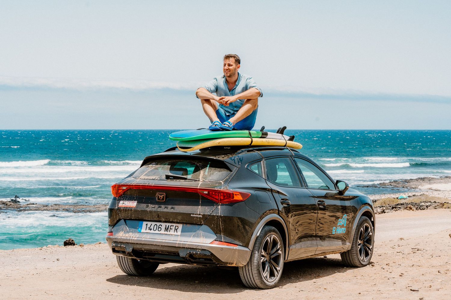 A luxury vehicle parked on sandy beach terrain with surfing equipment on the roof.