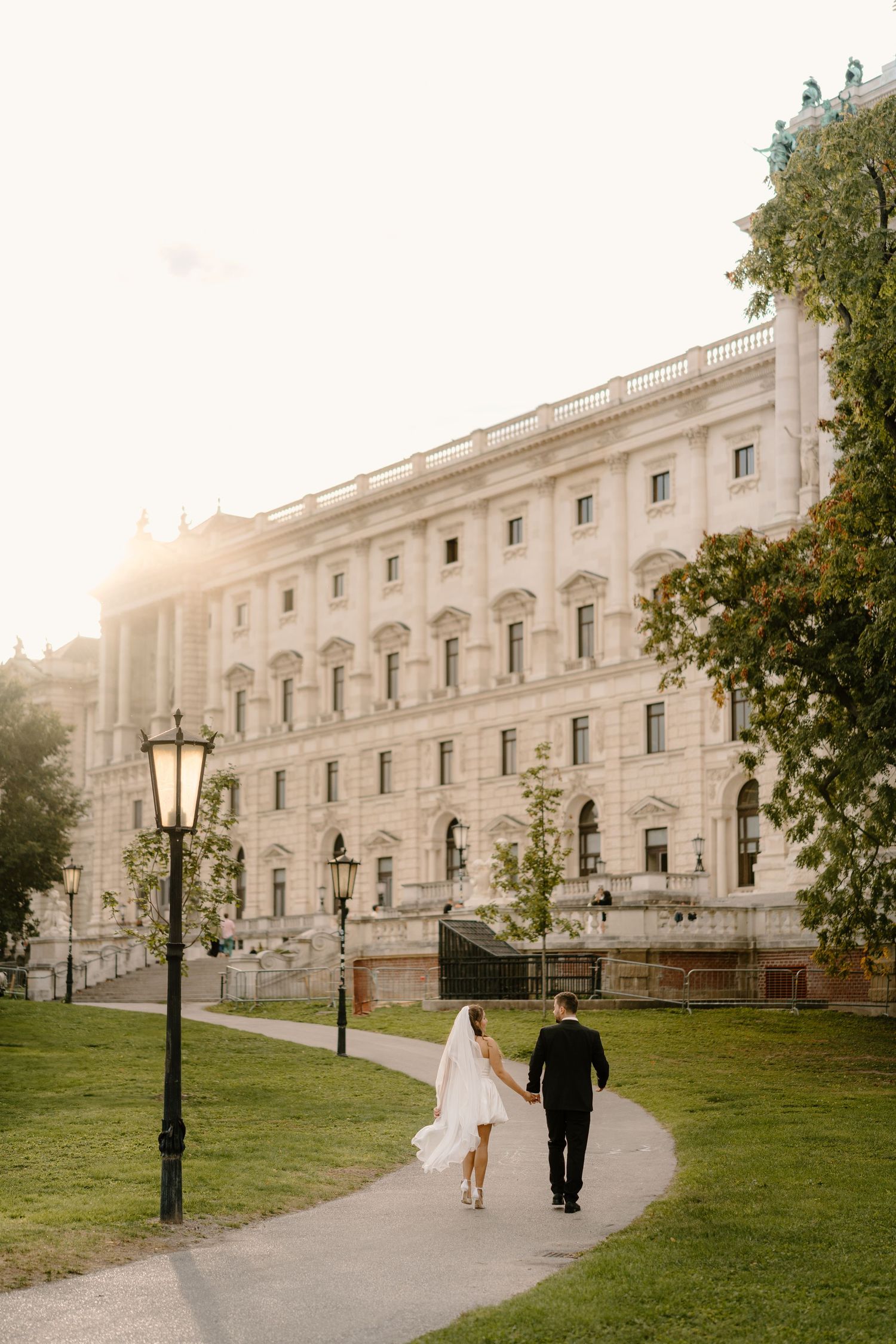 A couple walks along a curved path near a grand historic building with ornate architecture at sunset.