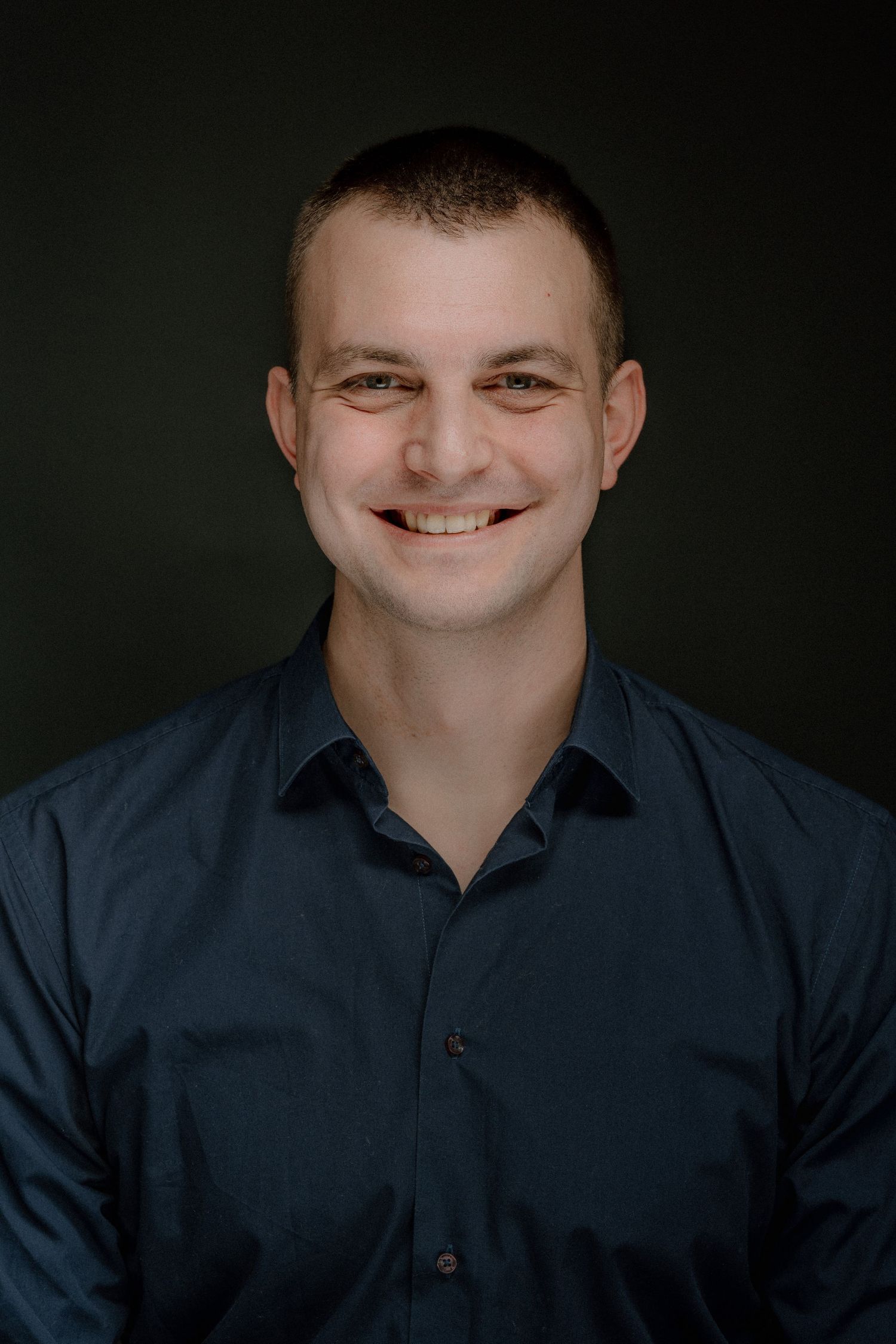 Smiling young man with short hair wearing a dark blue button-up shirt against a dark background.