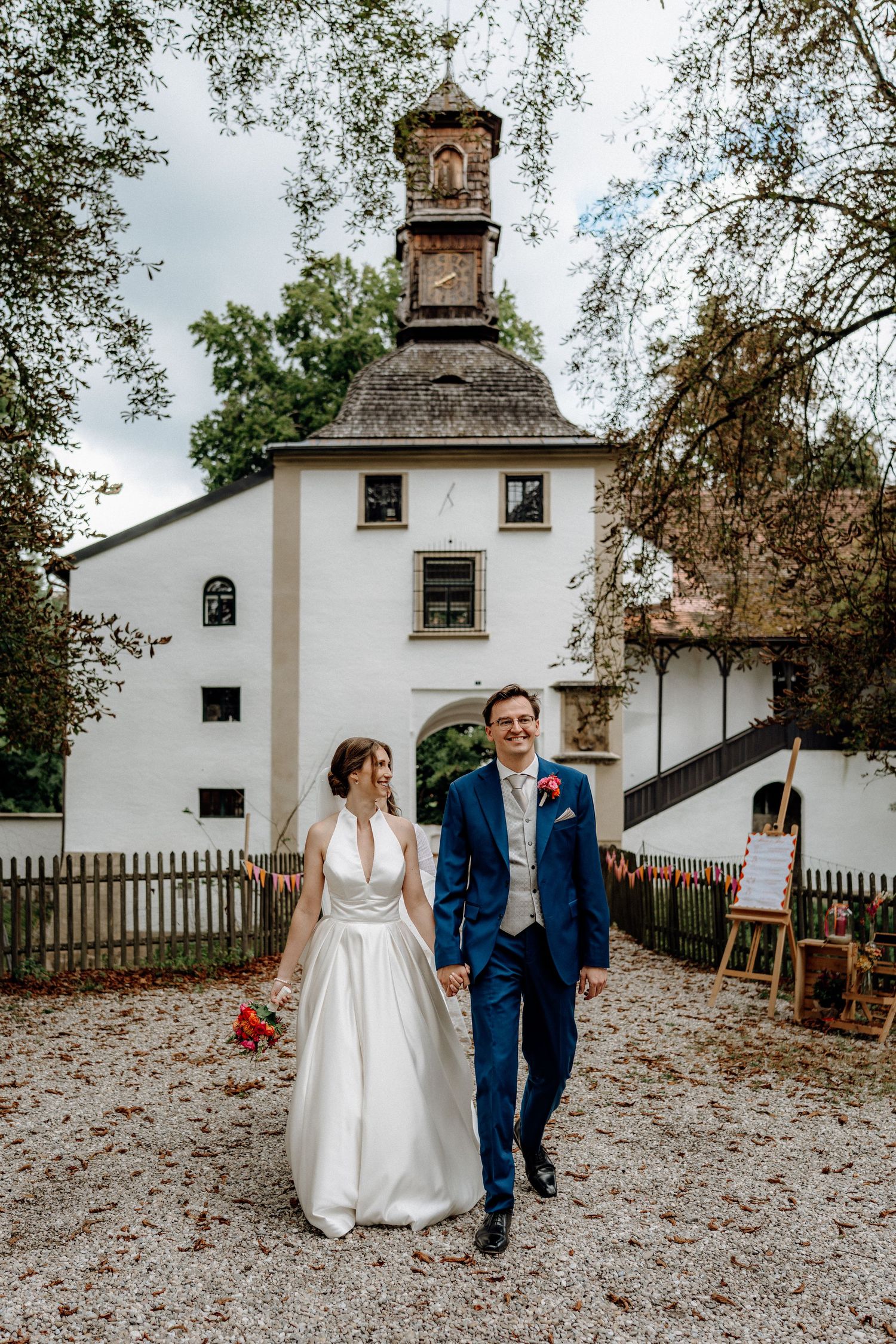 A couple walks together on a white gravel path outside a historic white building with a small tower.