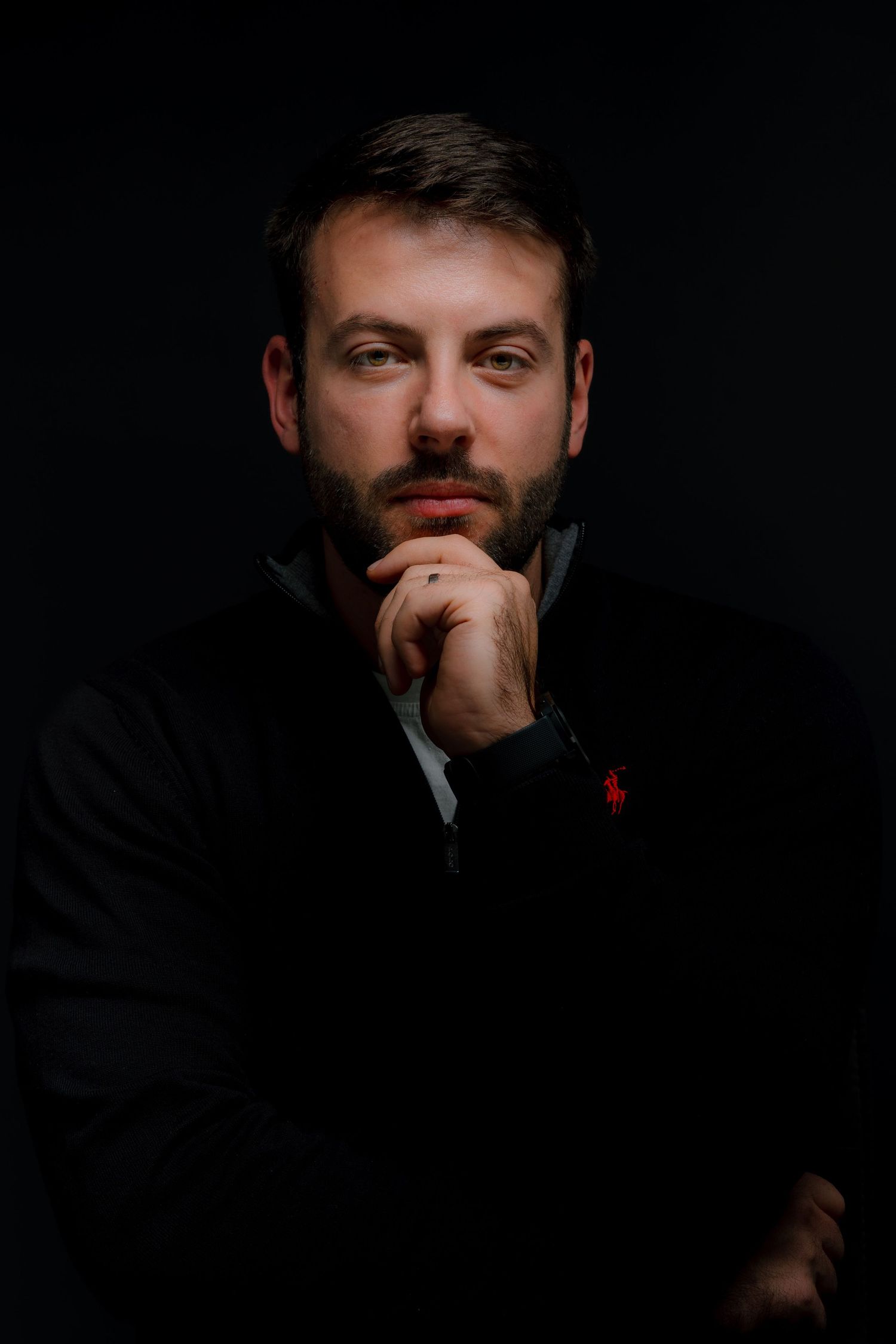 Thoughtful bearded man in black sweater poses with hand on chin against dark background in dramatic portrait.