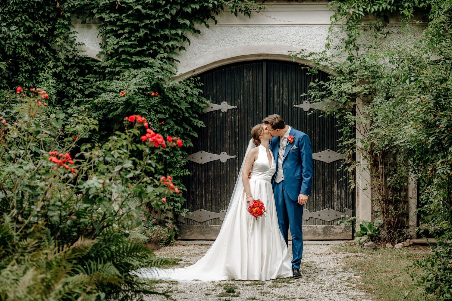 Wedding photography session in front of a rustic stone building with red flowers and greenery.
