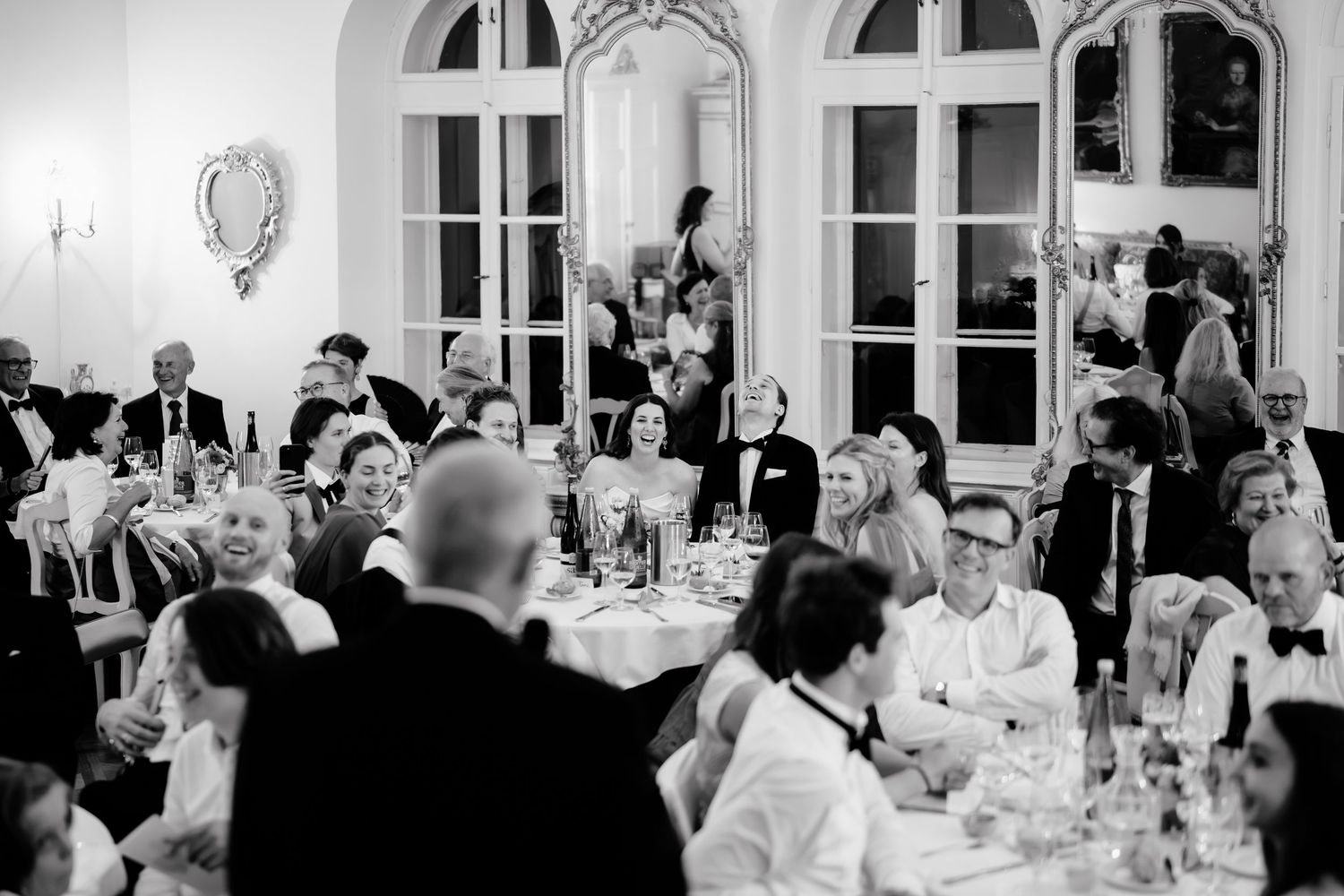 Black and white wedding reception photo showing elegantly dressed guests seated at tables in a grand ballroom.