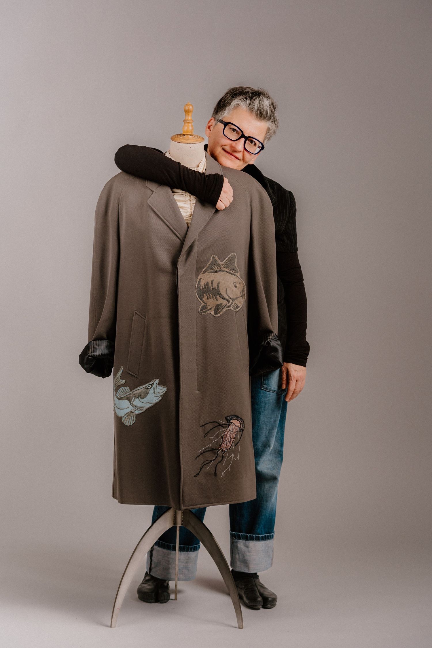 Older woman with glasses smiling beside a mannequin wearing an embroidered olive coat with fish and bird designs.