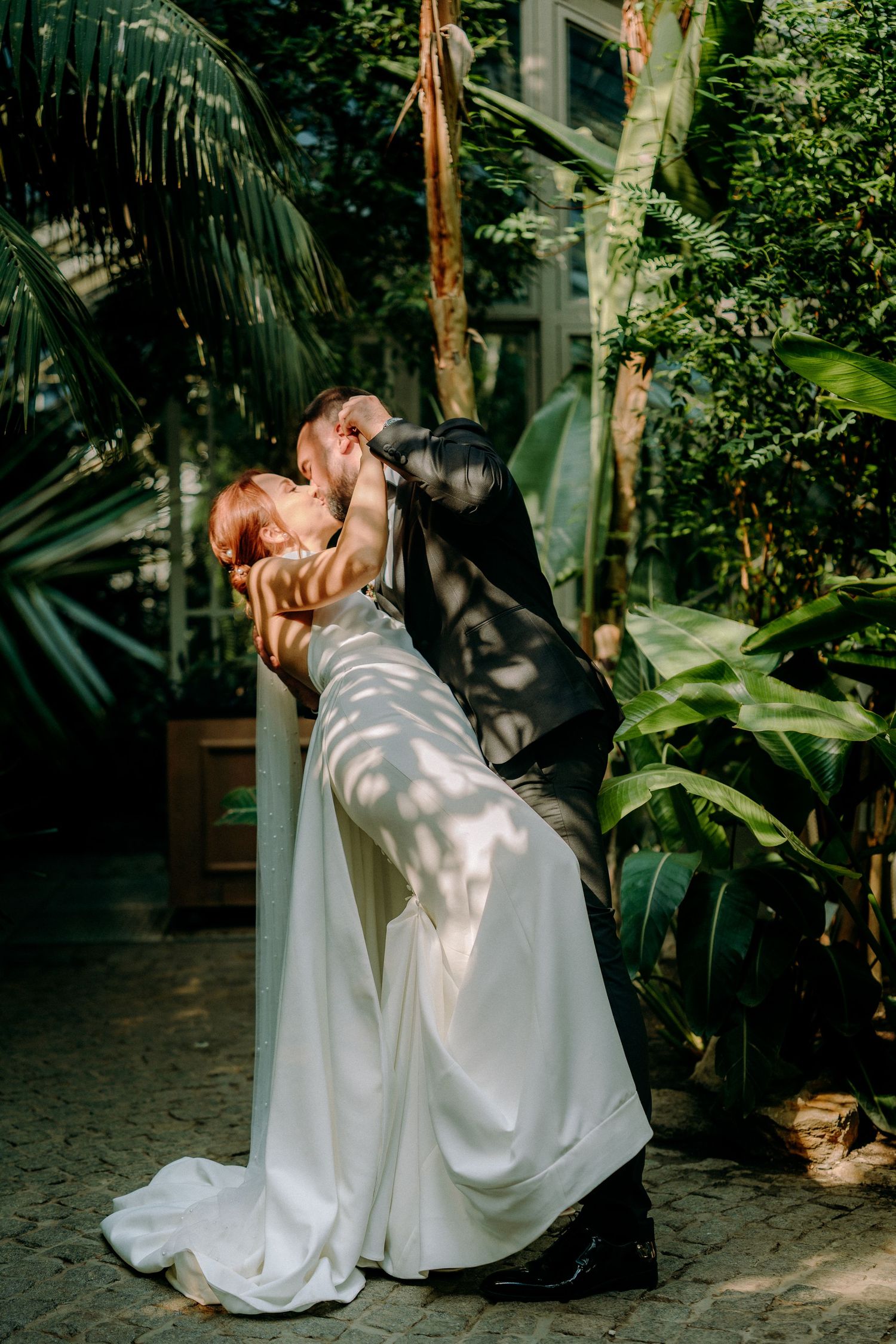 Wedding couple sharing intimate moments in a sunlit greenhouse surrounded by tropical foliage.