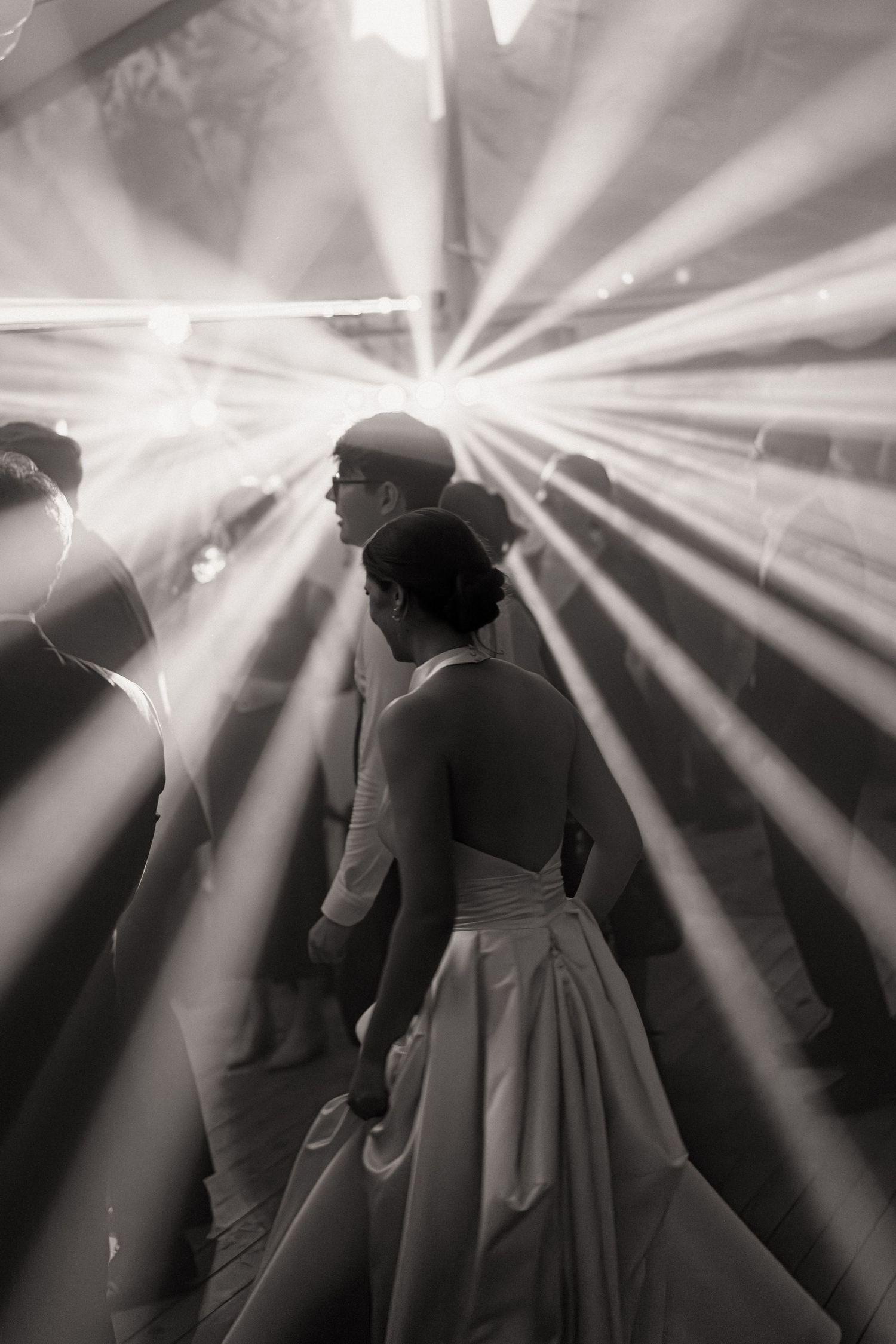 Black and white photo with dramatic light beams creating silhouettes at a wedding reception.