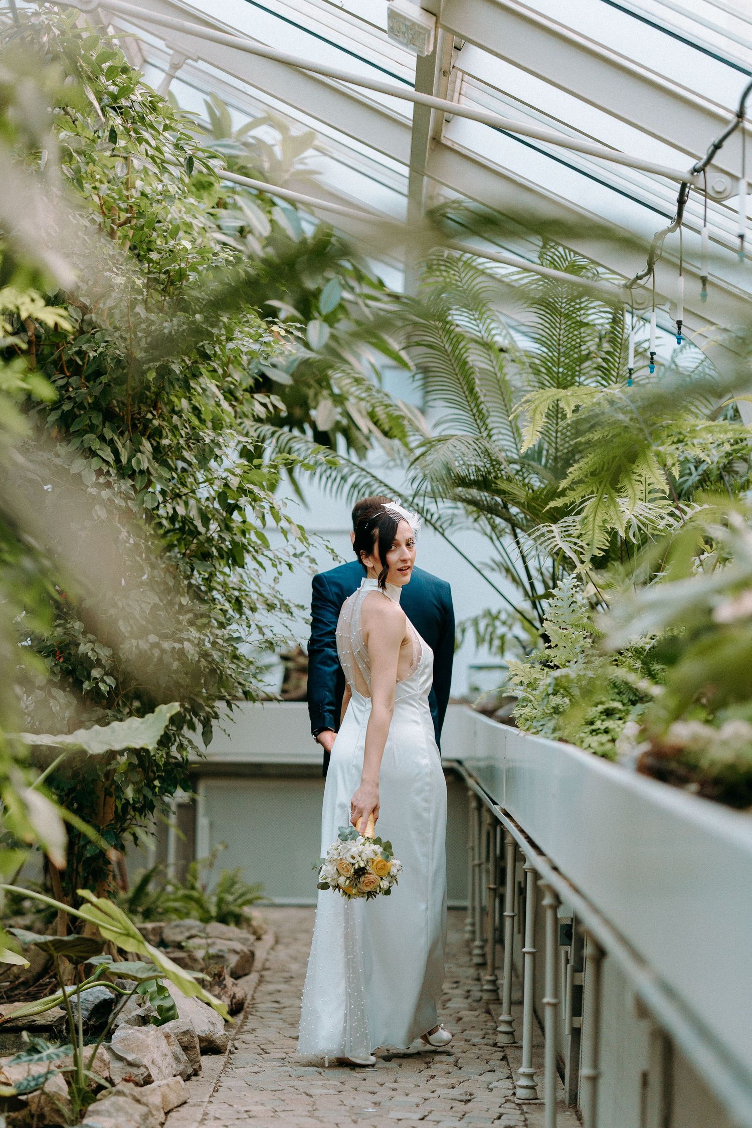 Bride and groom share a romantic moment in a lush greenhouse, bride holding a wildflower bouquet in a white gown.