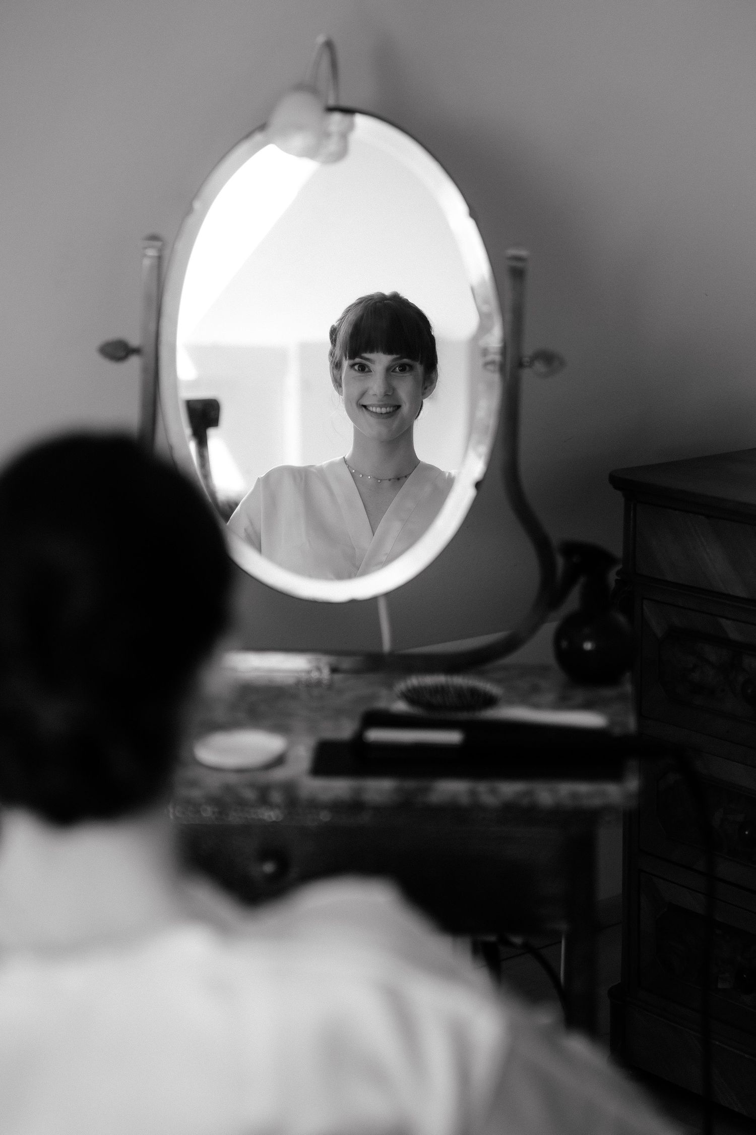 Black and white photo of a smiling woman reflected in an oval vanity mirror while sitting at a dressing table.