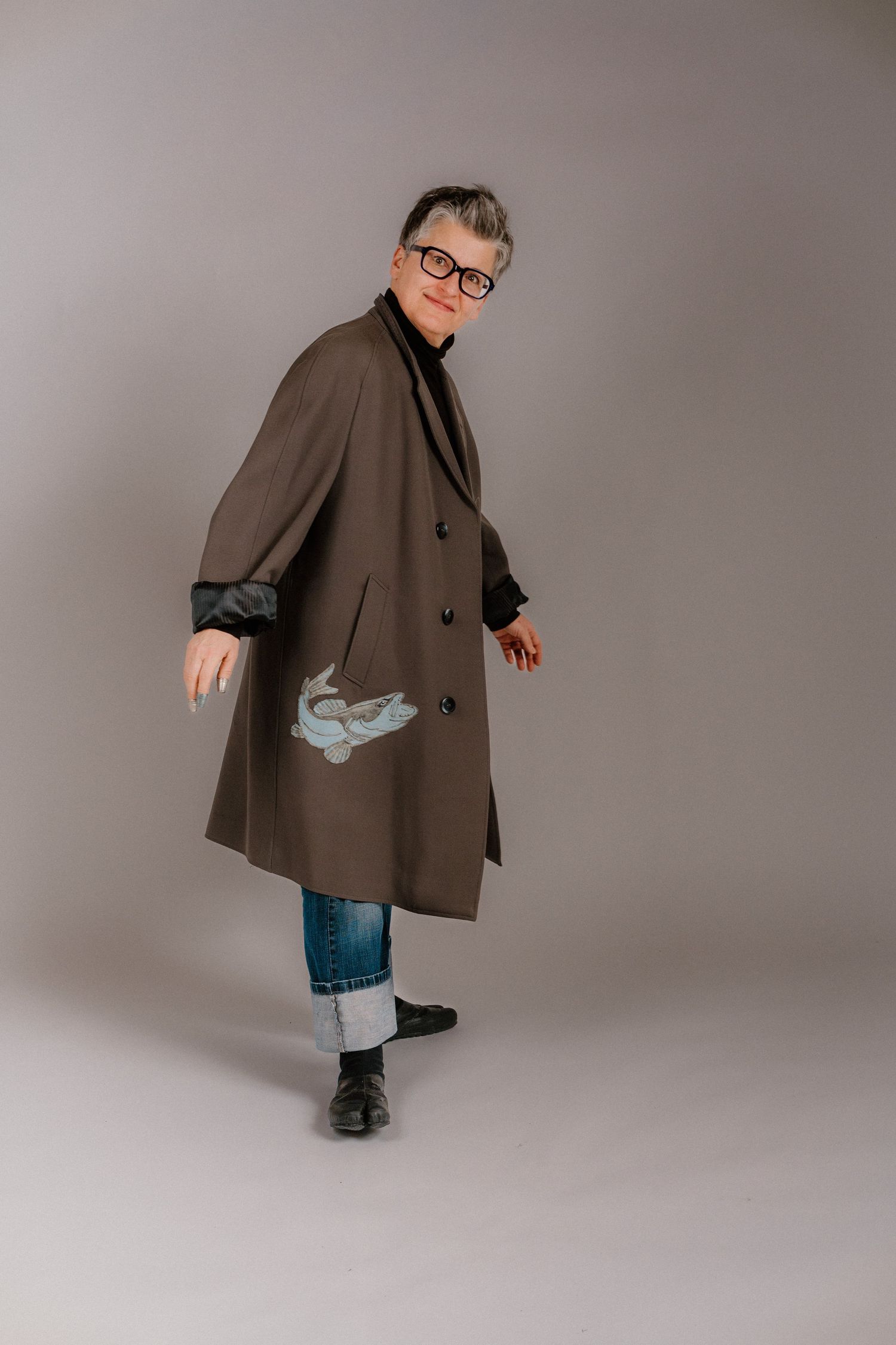 Woman in oversized brown coat with bird print, jeans, and glasses poses in studio.