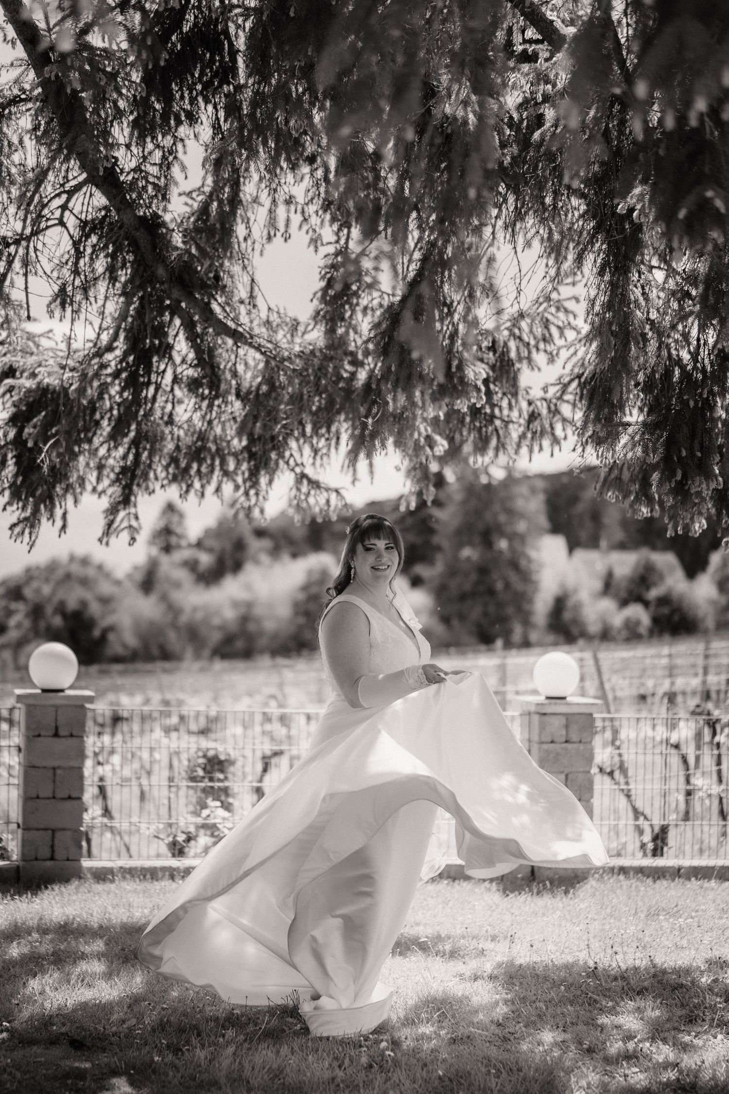 Black and white photo of a bride in a flowing white gown twirling under a pine tree in a garden.