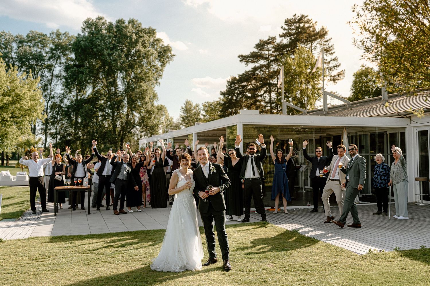 Bride and groom stand together outdoors as wedding guests celebrate behind them on a sunny day.
