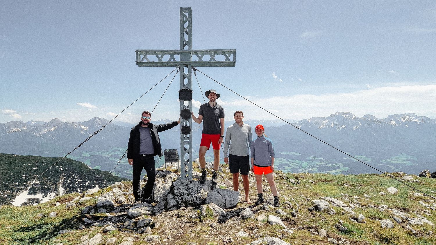 Group standing at summit cross on mountain peak with snow-capped mountains in background.